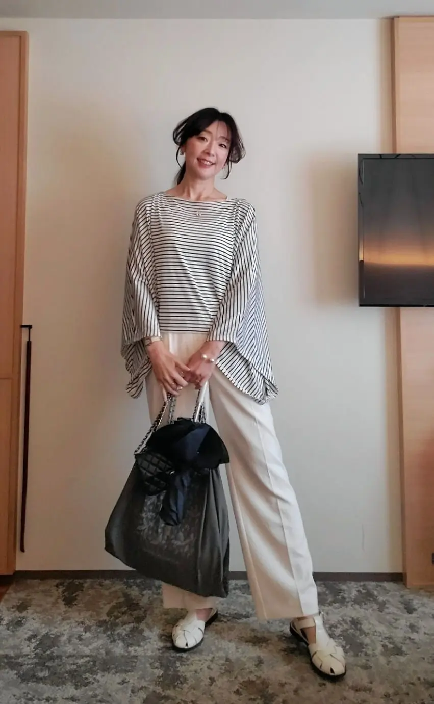 黒と白のストライプのゆったりとしたトップス、白いワイドパンツを着用した女性。黒いバッグを持ち、白い靴を履いている。背景にはテレビと木製の壁がある。
