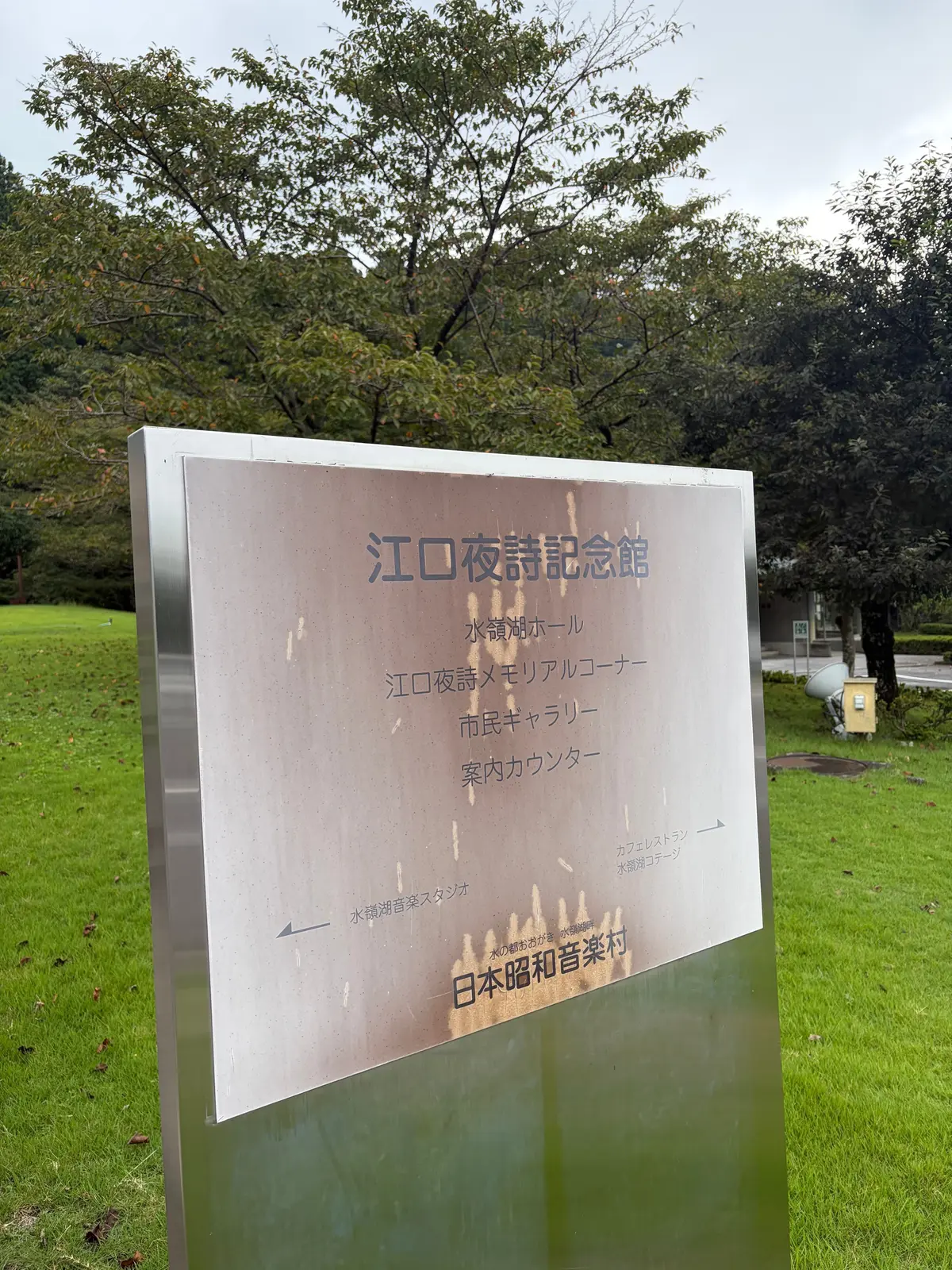 ステンレス製の標識で、青色の文字で「江口夜話記念館」と大きく表示されている。水鏡ホール、江口夜話メモリアルコーナー、市民ギャラリー、案内カウンターなどの施設名が記載され、下部には「日本昭和音楽村」の文字がある。