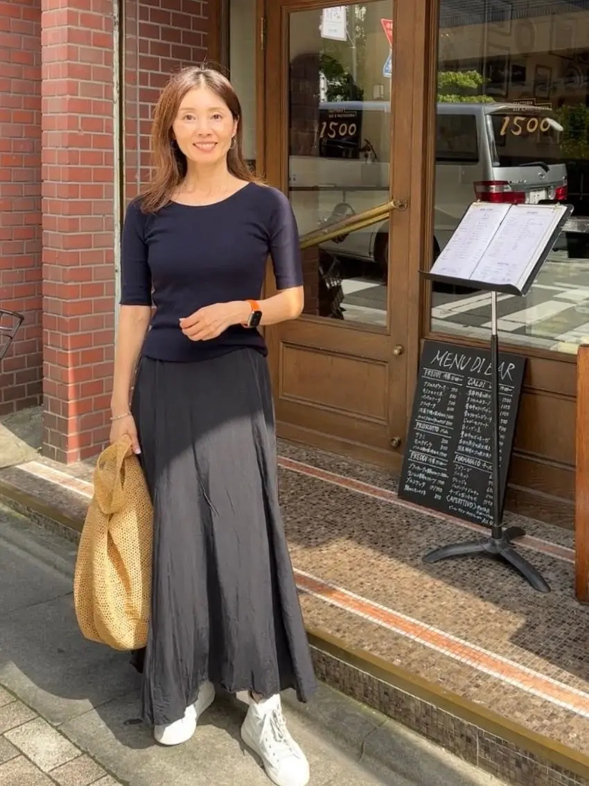 紺色のトップスと黒いロングスカートを着用した女性が、カフェの前で立っている。右手にベージュのトートバッグを持ち、白いスニーカーを履いている。背景には、メニュー立てと黒い車が見える。