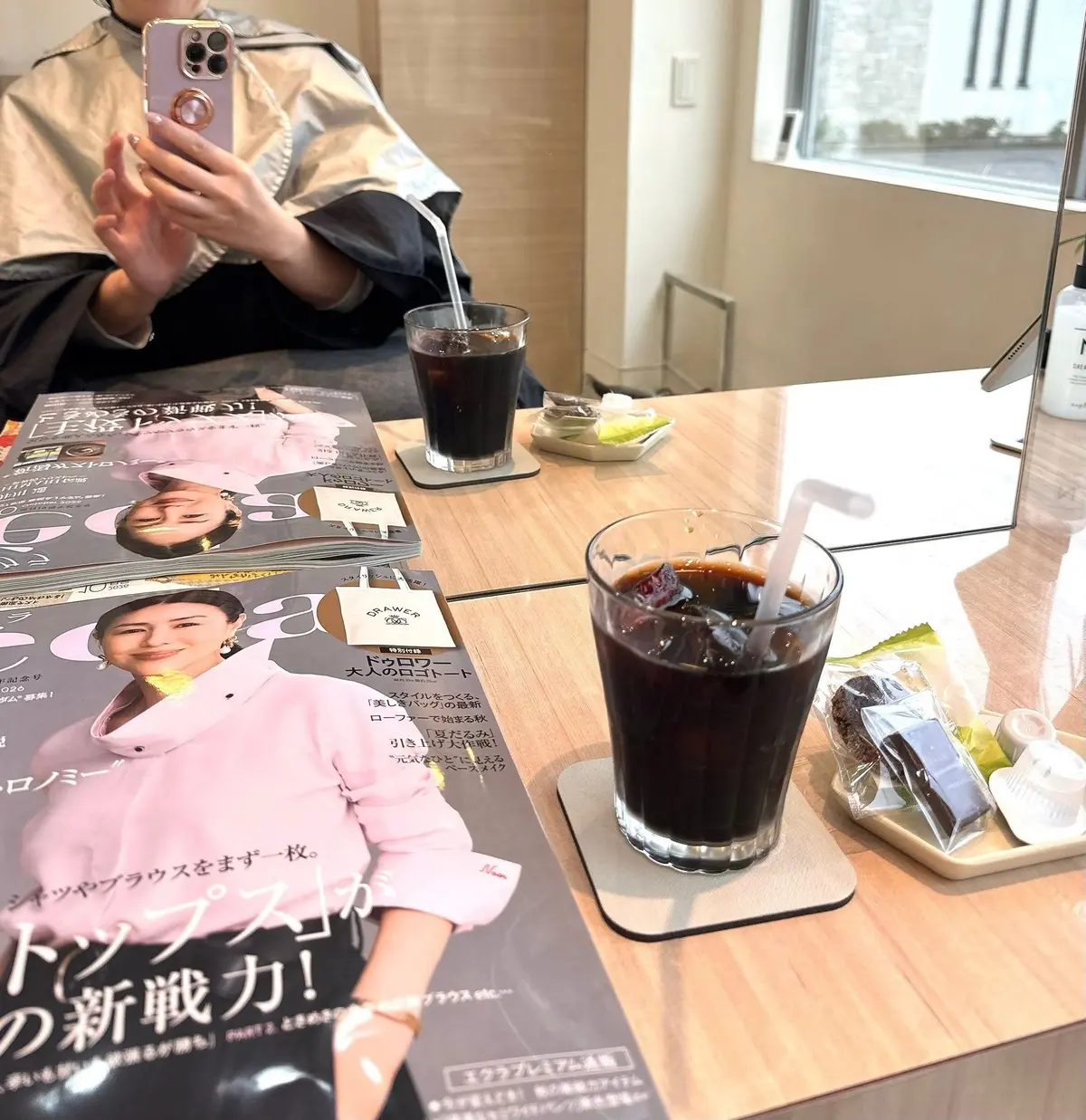 電車の座席でスマートフォンを見ている女性。テーブルにはファッション誌と、グラスに入ったサラダが置かれている。背景には窓から見える景色。