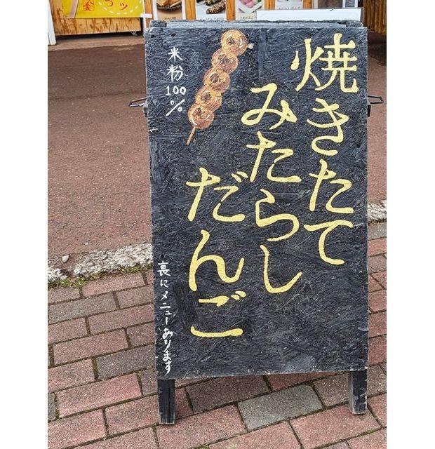 黒板に手書きで「焼きたて みたらしだんご」と書かれた看板。上部には「米粉100%」の文字と、イラストが描かれている。下部には小さく「裏にはメニューあります」と記載。