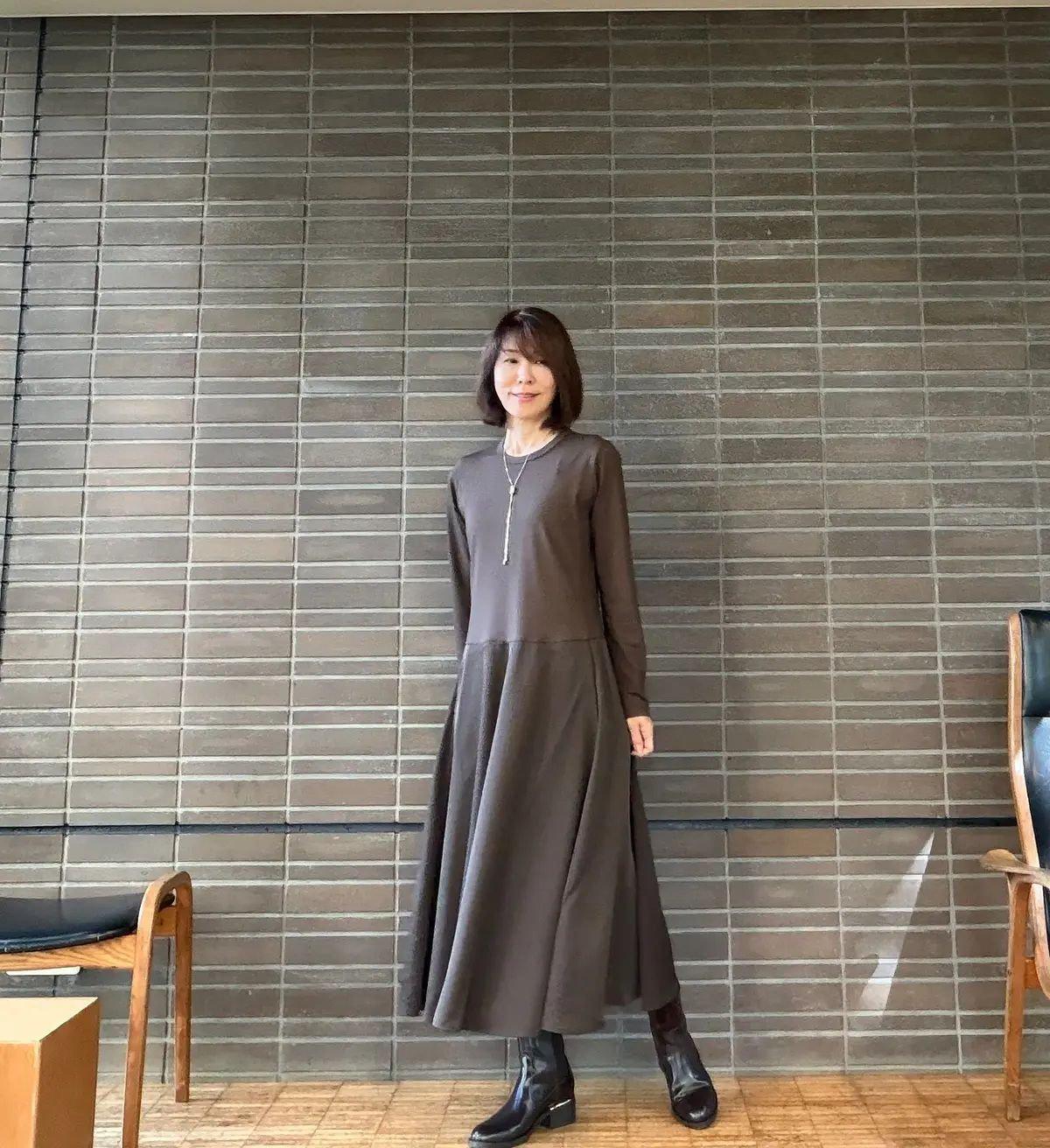 茶色のロングワンピースを着た女性が、グレーの壁の前で立っている全身写真。黒いブーツを履き、シルバーのネックレスを着用。髪は肩まで切りそろえられている。