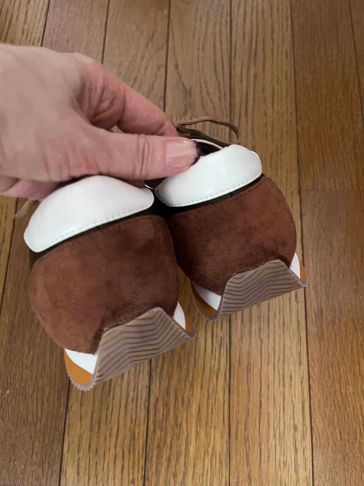 茶色のスエード素材の靴のソール部分が2つ並んでいる。白い縁取りがあり、靴紐の先端が白く見えている。背景は木製の床。