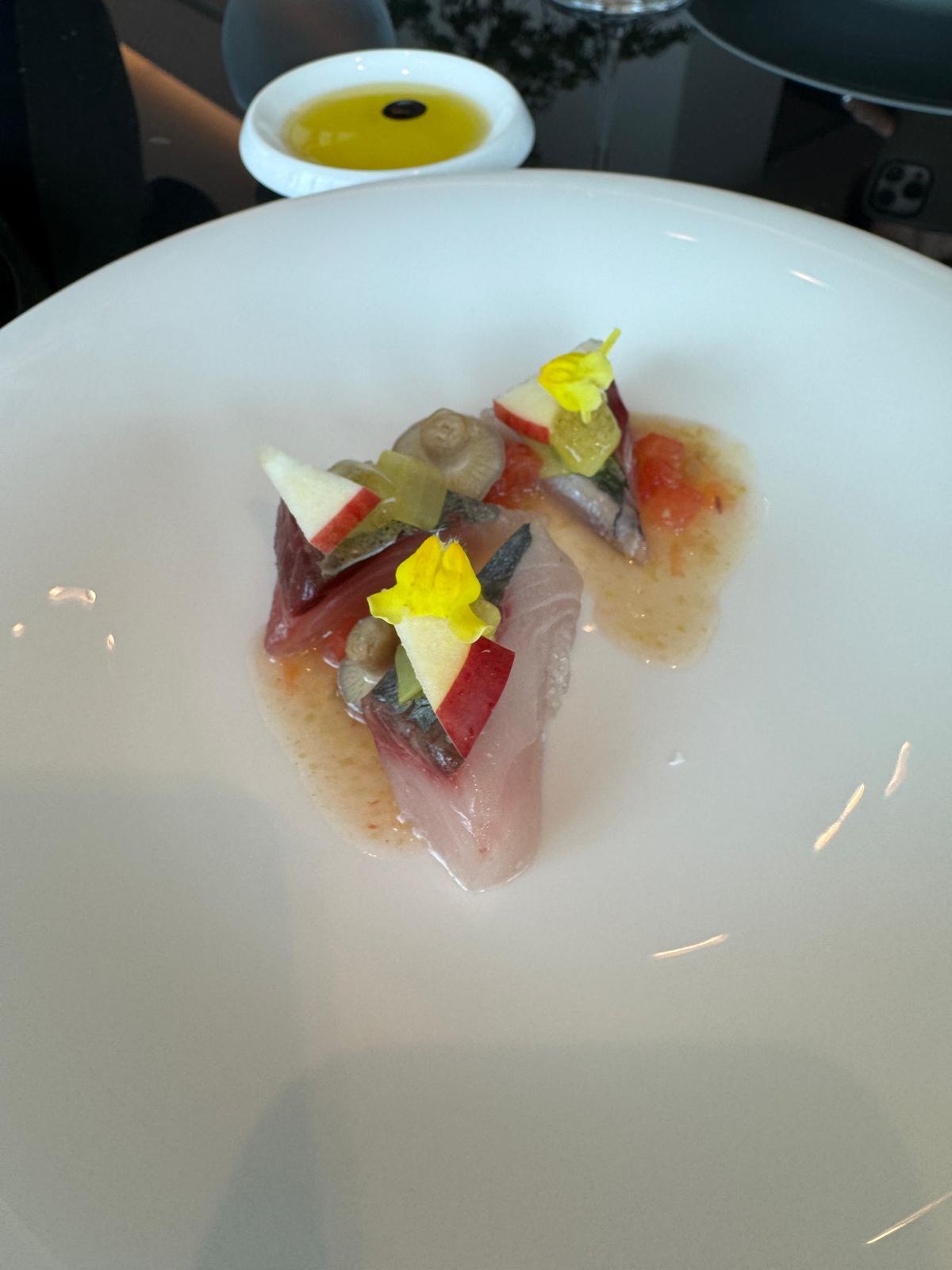 白い皿に盛り付けられた、赤身の魚と赤い果肉、ピクルス、黄色い花を添えた料理。皿の周りにはオレンジ色のソースが点在している。背景には小さな器に入った液体と、黒い脚を持つテーブルスタンドが見える。