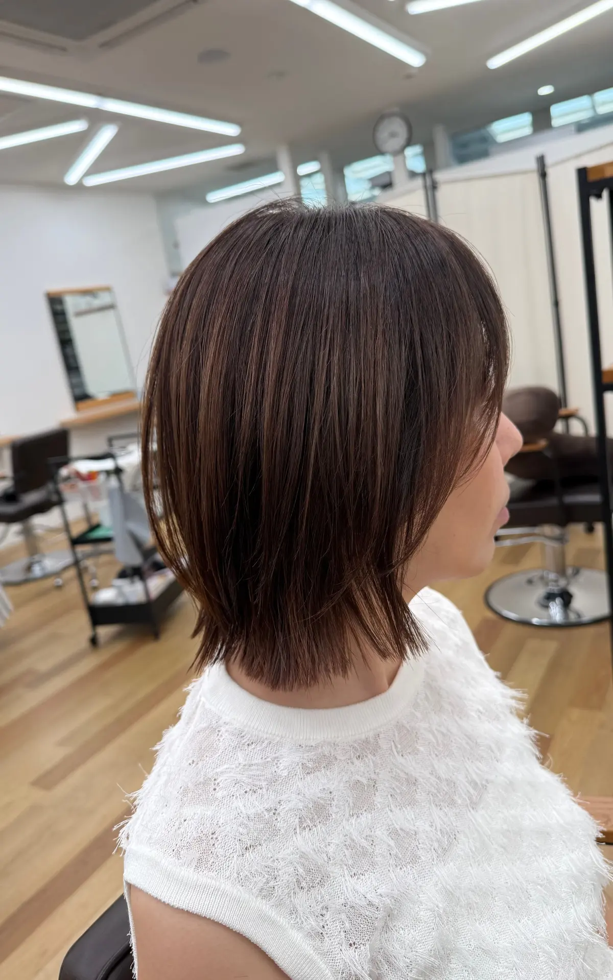 茶色のボブヘアの女性が、白いレースのトップスを着て座っている横顔。背景には美容室の設備が見える。