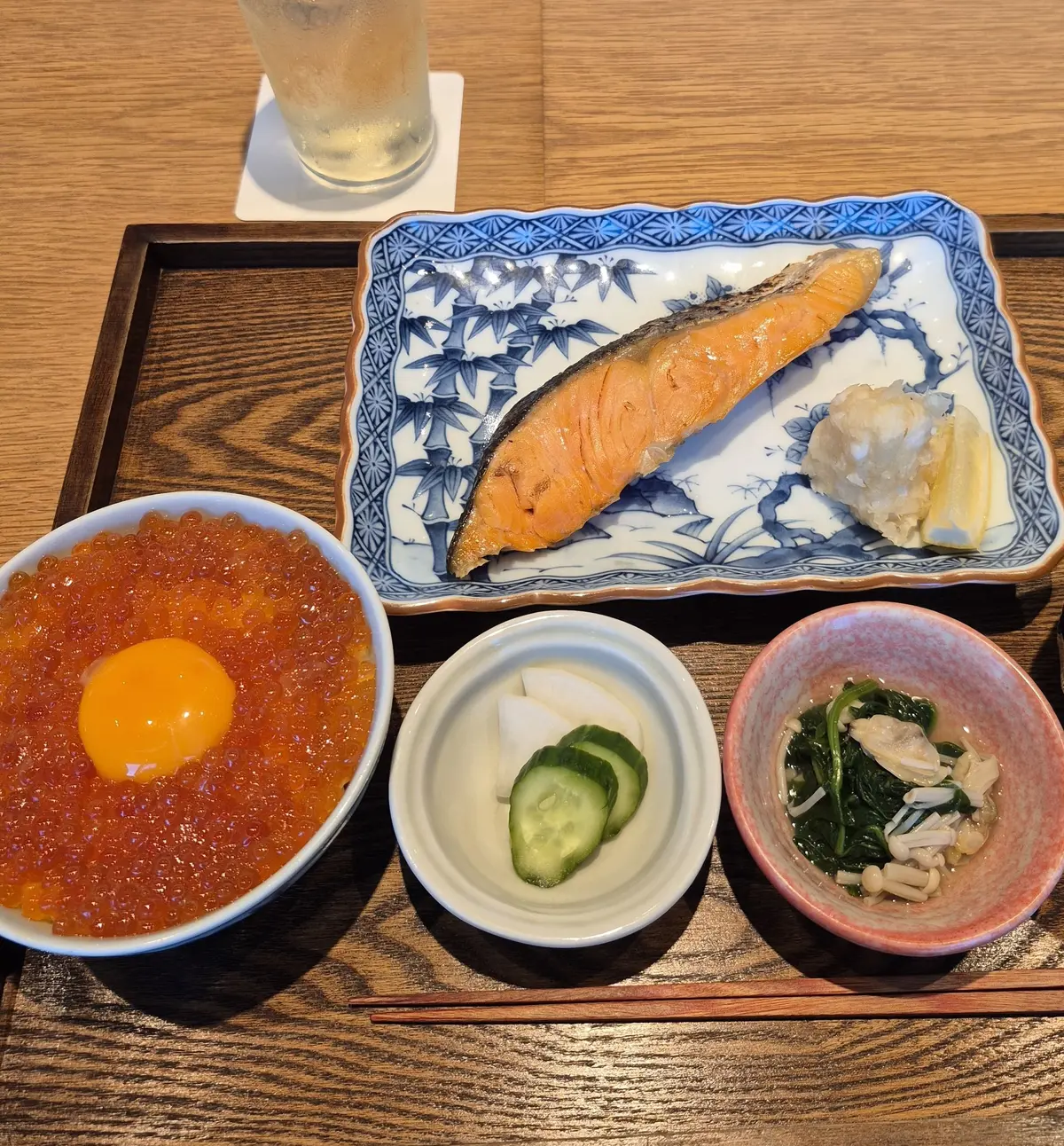 木製のトレイに盛り付けられた和食。鮭の塩焼き、いくらと卵の丼、お新香、ほうれん草のおひたし。グラスに入った飲み物とメニューも写っている。