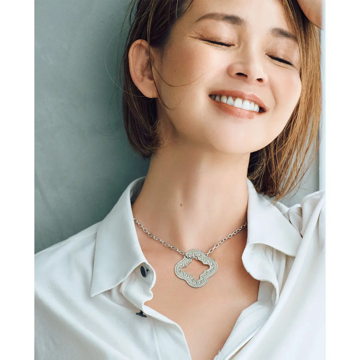 笑顔の女性が目を閉じ、白いシャツを着用。銀色のネックレスを身につけ、背景は淡いブルー。