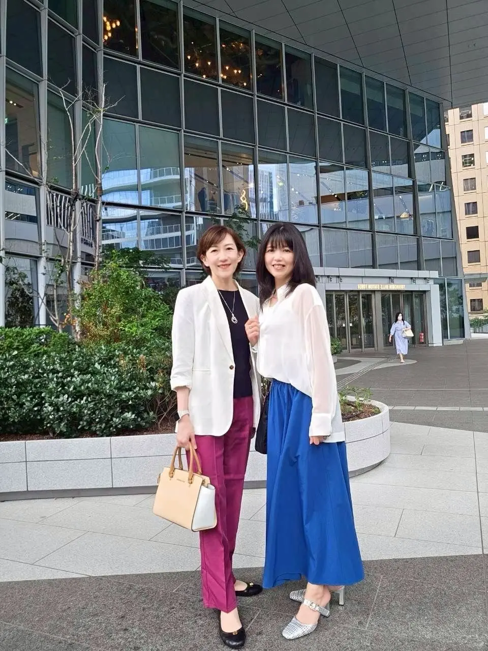 二人の女性が笑顔で並んで立っている。左の女性は白のブレザーと紫のパンツ、右の女性は白のブラウスと青のロングスカートを着用。左の女性はベージュのトートバッグを手に持っている。背景にはガラス張りの建物と低木が並ぶ。