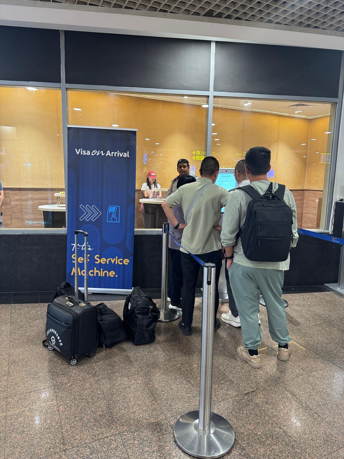 空港の「Visa on Arrival」カウンター前で、3人の男性が手続き待ち。男性はそれぞれスーツケースを持ち、スマートフォンを見ている。背景には窓から光が差し込み、黄色の服を着たスタッフがいる。