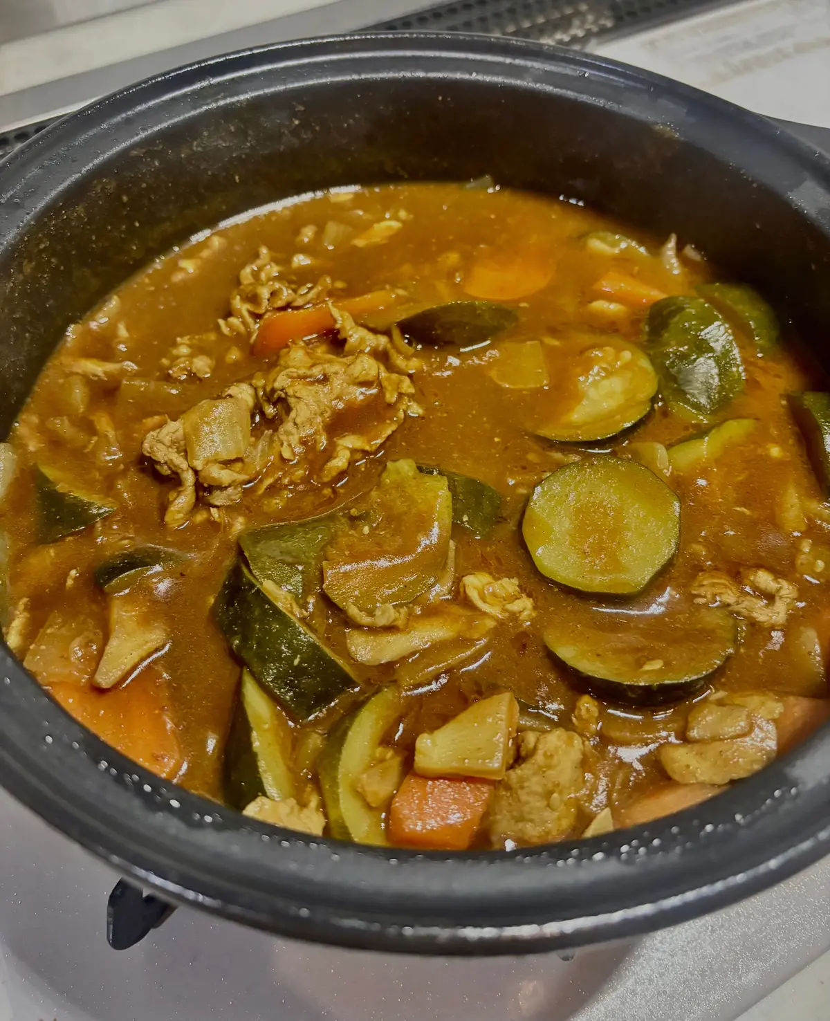 黒い鍋に入った、茶色いカレー。豚肉、人参、ズッキーニがごろごろと入っている。表面はとろみがあり、湯気が出ている。