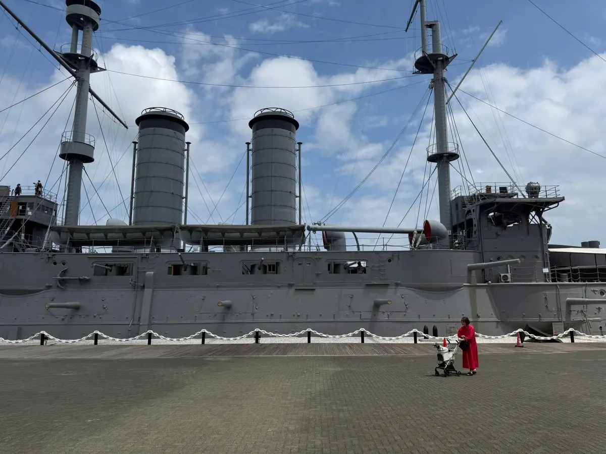 灰色で巨大な戦艦が、青空と雲の下に停泊している。船体には複数の煙突と、細い鉄の柵が設置されている。手前に女性が赤いコートを着て、ベビーカーを押している。