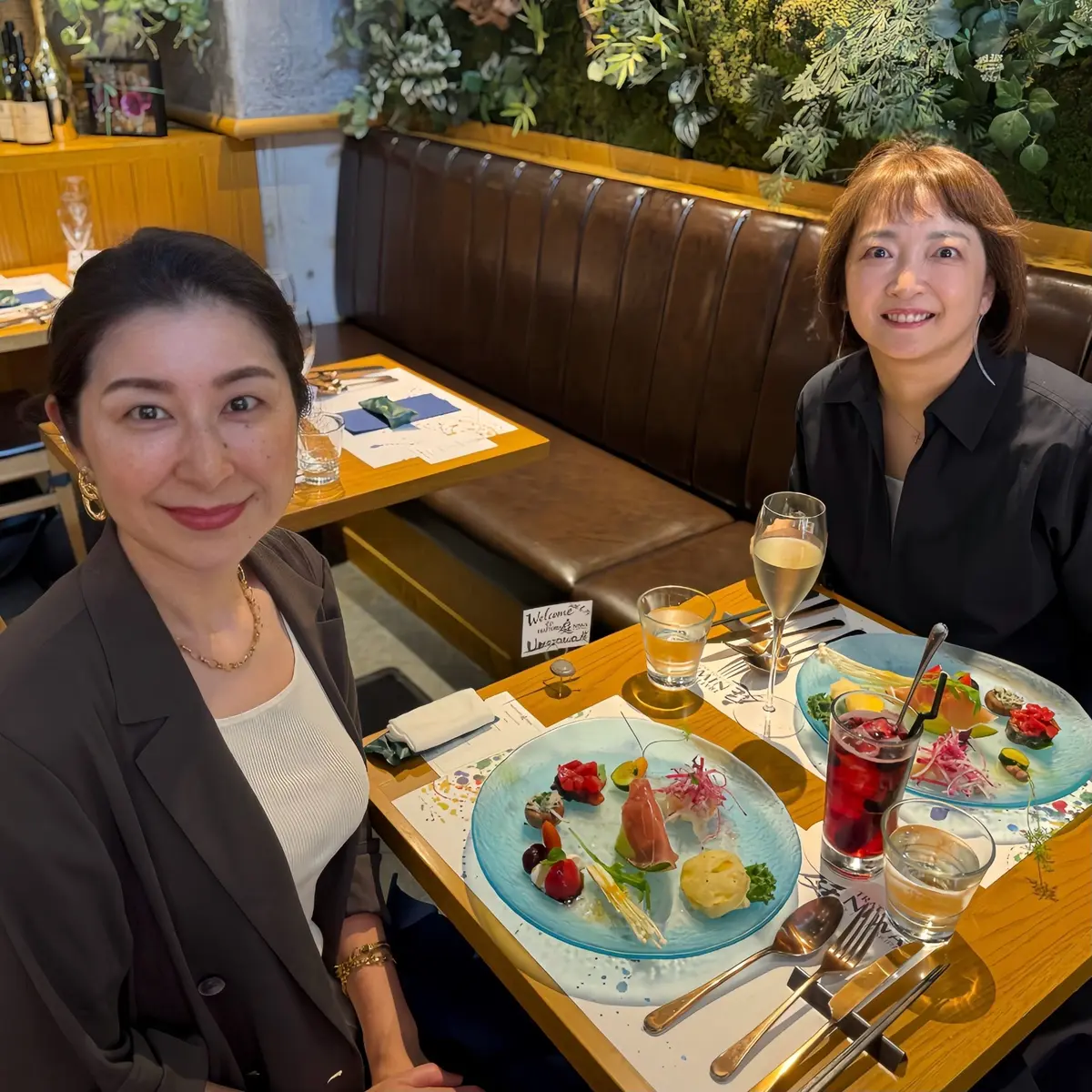 表参道レストラン、イタリアンレストラン庭、木製テーブルに女性二人が座り、前に料理とドリンクが並んでいる。左の女性は黒のブレザージャケットを着て、右の女性は黒のシャツを着ている。