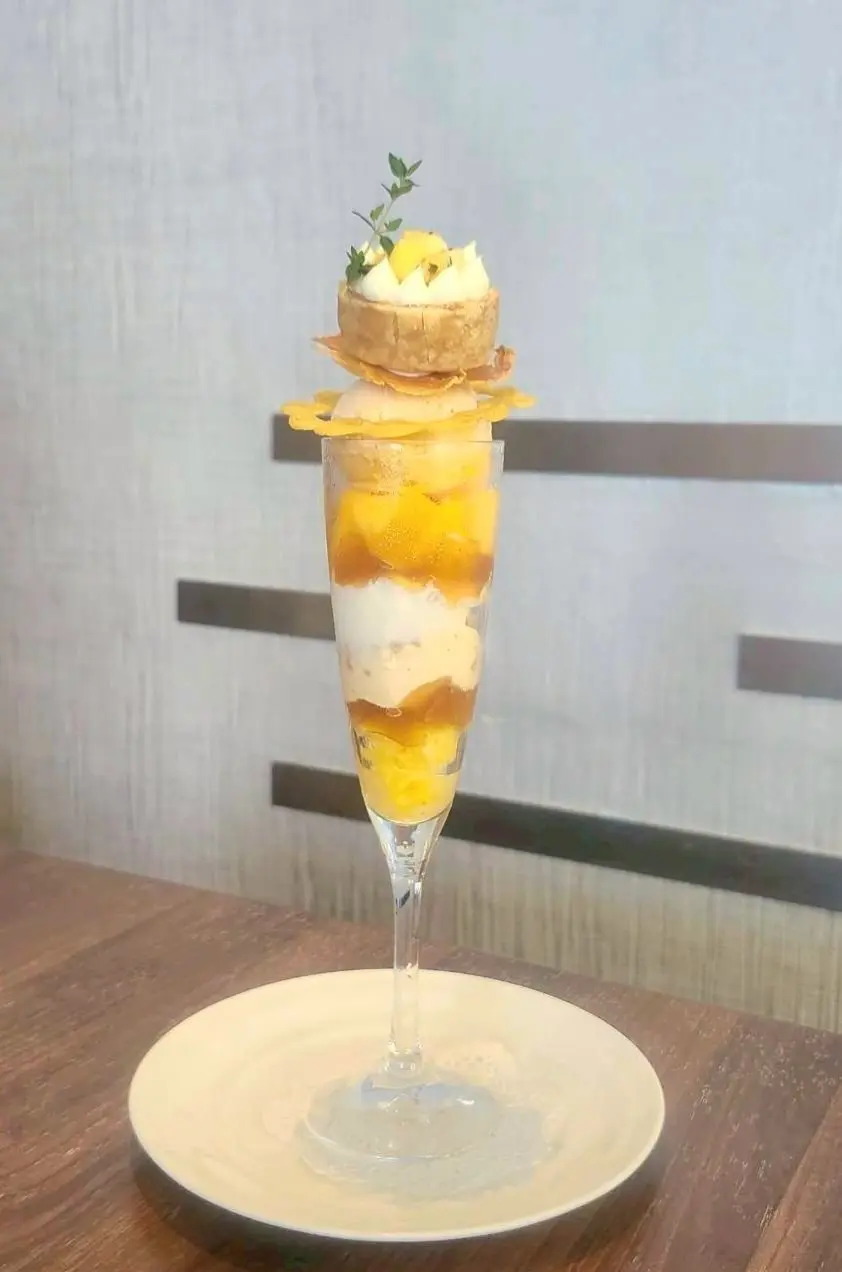 ガラスカップに層になったフルーツとクリーム、クラストが乗せられたデザートで、トップには小さなハーブの葉が飾られている