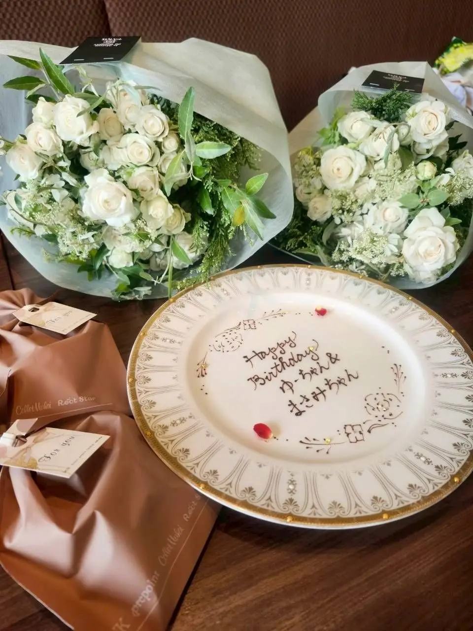 白いバラとグリーンの葉で構成された2つの花束と、金の縁取りと「Happy Birthday」の文字が書かれた白いプレートが木製のテーブルに置かれている。