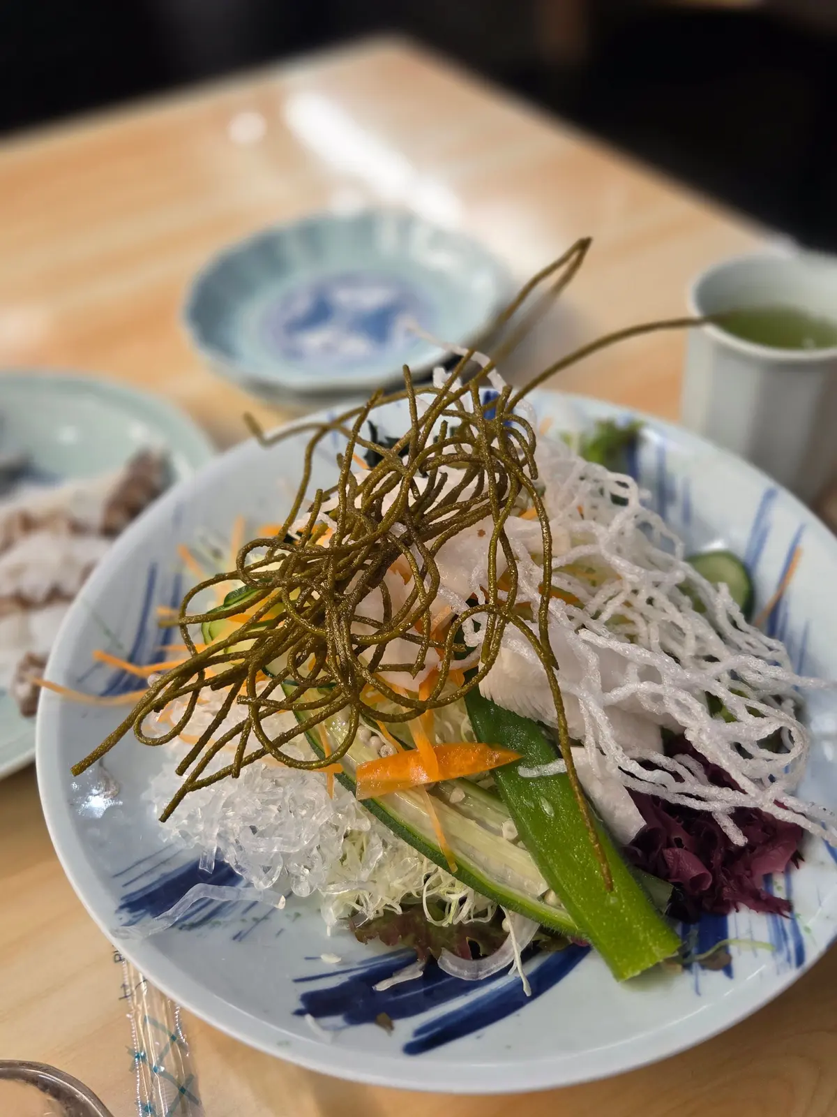 青い模様の皿に盛り付けられた、緑色の細長い麺と白く繊細な麺、薄切りキュウリ、赤と紫の野菜、オレンジ色の飾り切りが配置された料理。背景には茶碗と他の皿が見える。