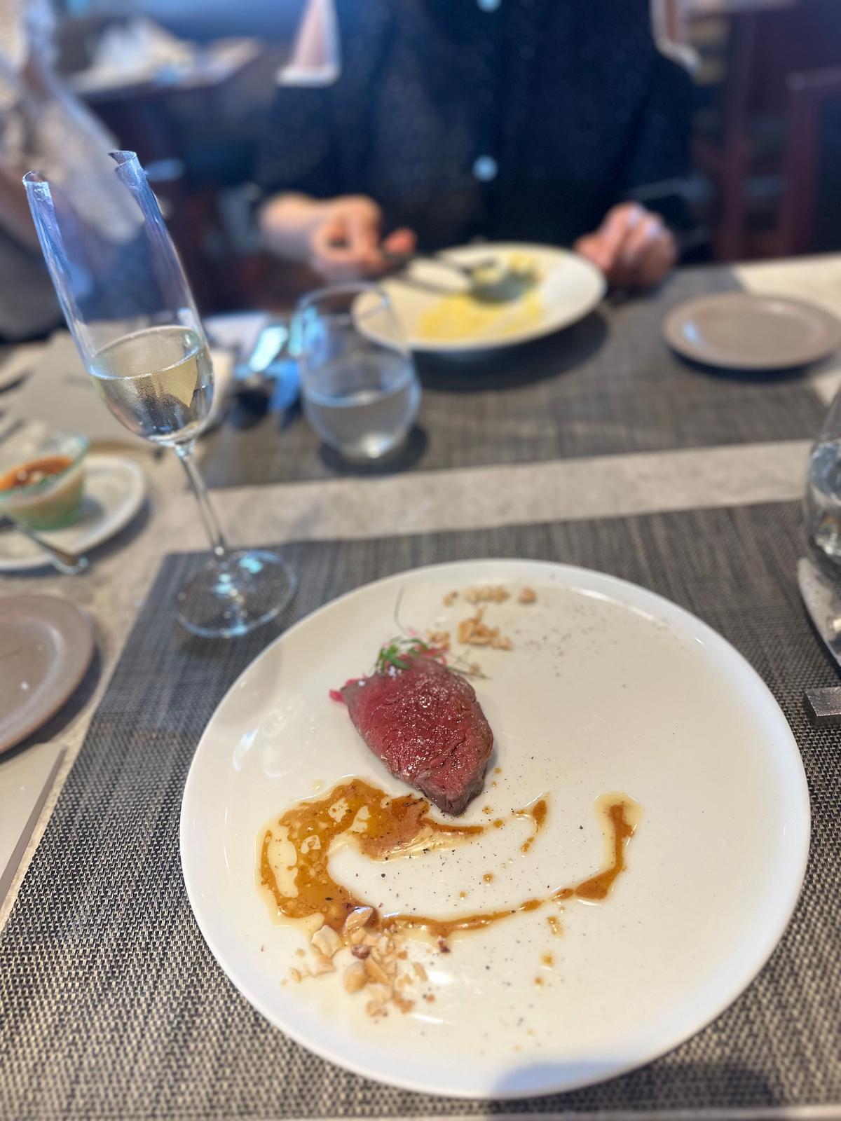 白磁の皿に盛り付けられた赤身の肉料理。ソースと砕いたナッツ、ドライハーブが添えられている。グラスに入った白ワインと、他の料理が背景に見える。