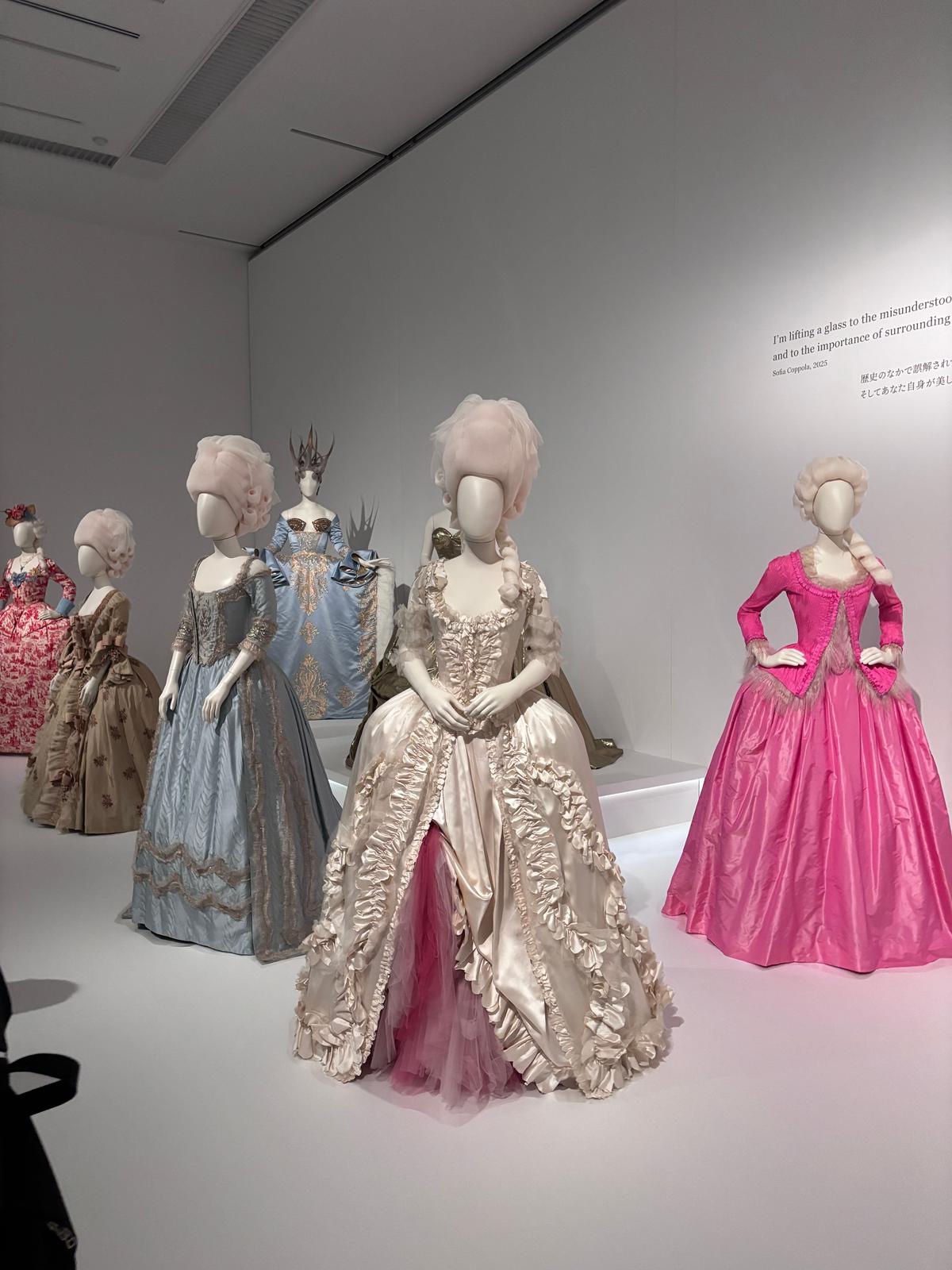 展示ケースに並んだ、18世紀風の豪華なドレスが7着。白、水色、ピンク、ゴールドなどの色合いで、フリルやリボン、レースなどの装飾が施されている。マネキンには、当時の髪型を模したウィッグが被せられている。