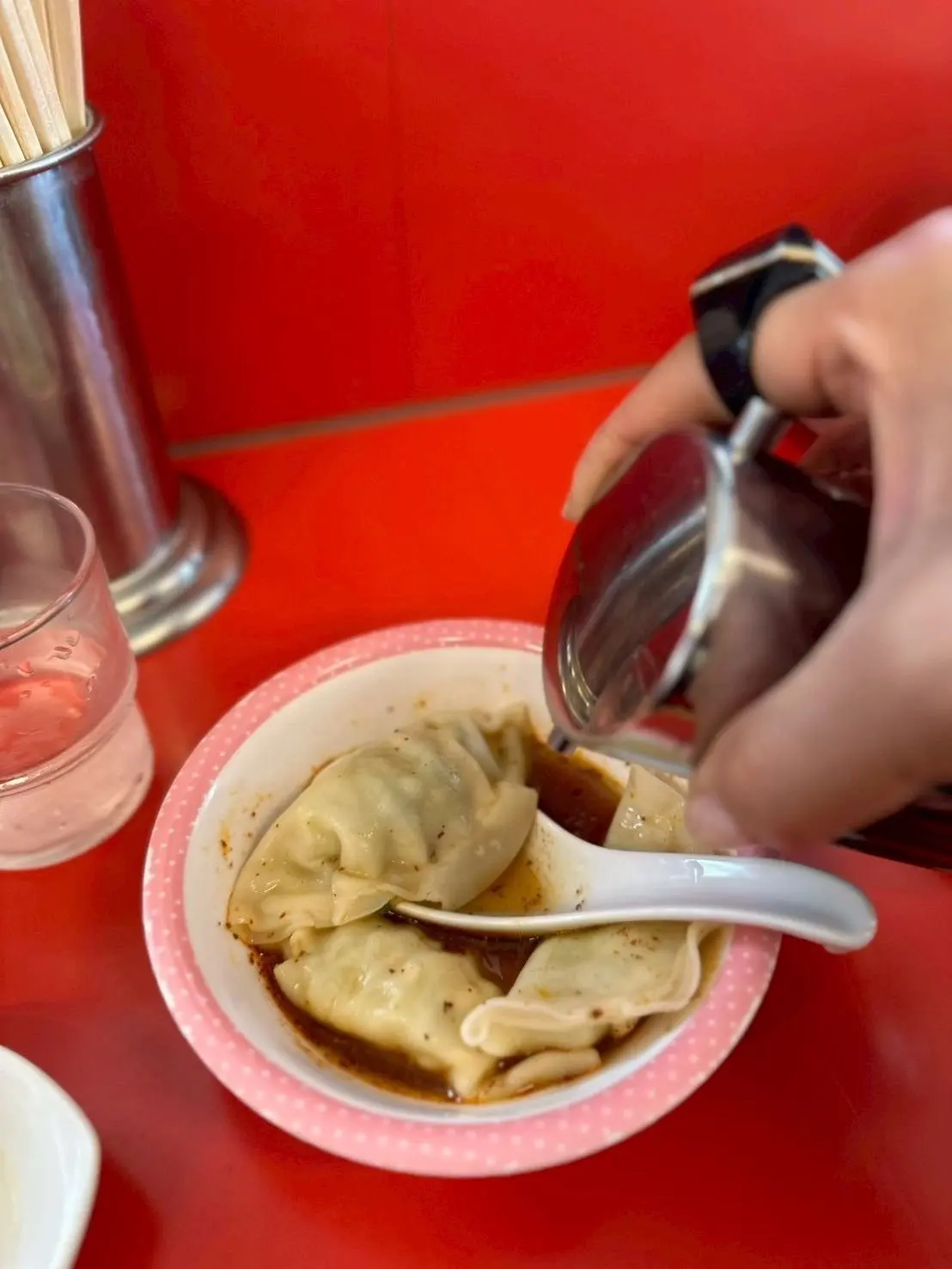 赤いテーブルに置かれた白い縁取りのボウルに、ジュワッとした餃子が並び、茶色のソースが注がれている。手はソースをスプーンで餃子にかけている。