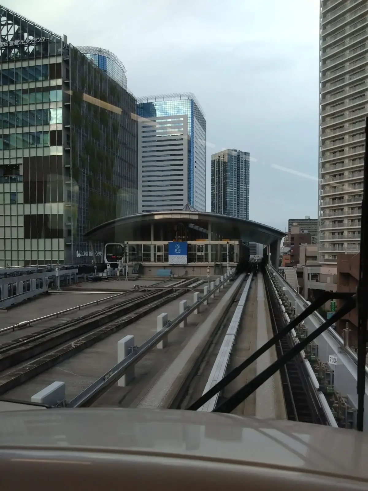 高層ビル群の間を走る、モノレールの駅の風景。駅舎は白を基調としたモダンなデザインで、モノレールが乗り入れ、駅の案内板が見える。背景には、ガラス張りの高層ビルが立ち並んでいる。