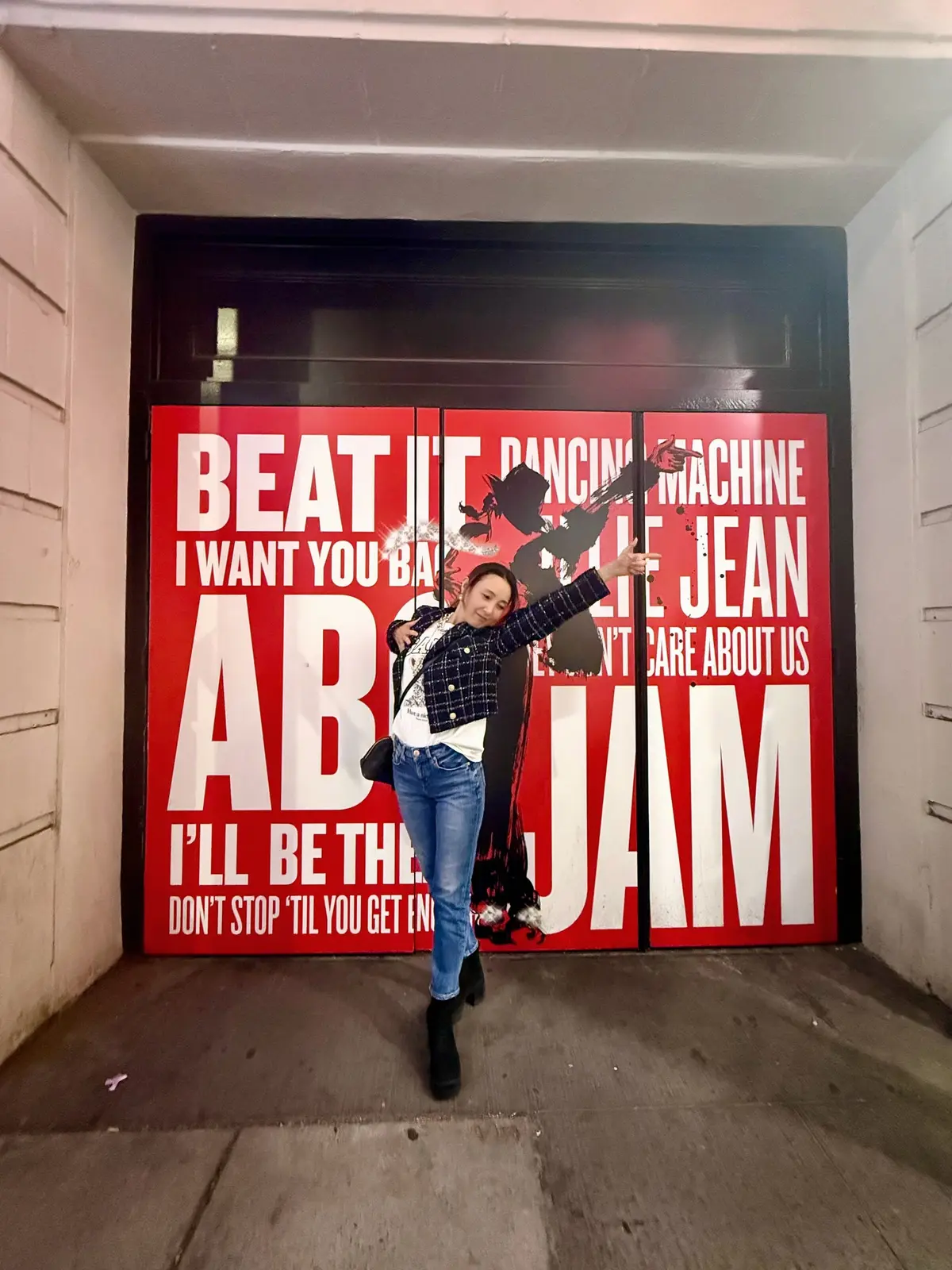 若者が赤い壁に貼られた「BEAT IT DANCING MACHINE」などの文字入りポスターの前で、両手を広げてポーズをとっている。青いチェック柄のジャケットとジーンズを着ている。