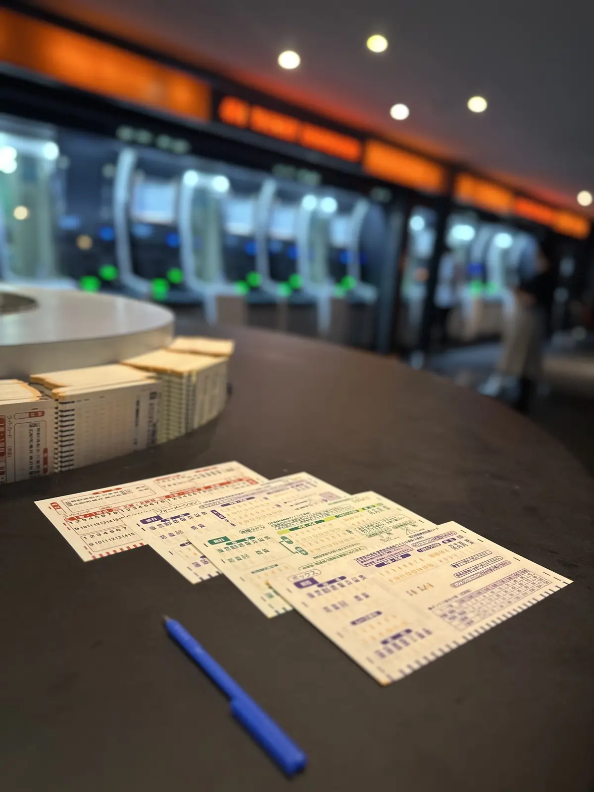 駅の窓口のカウンターに、切符や時刻表が置かれ、青いペンが添えられている。背景には自動改札機が見える。