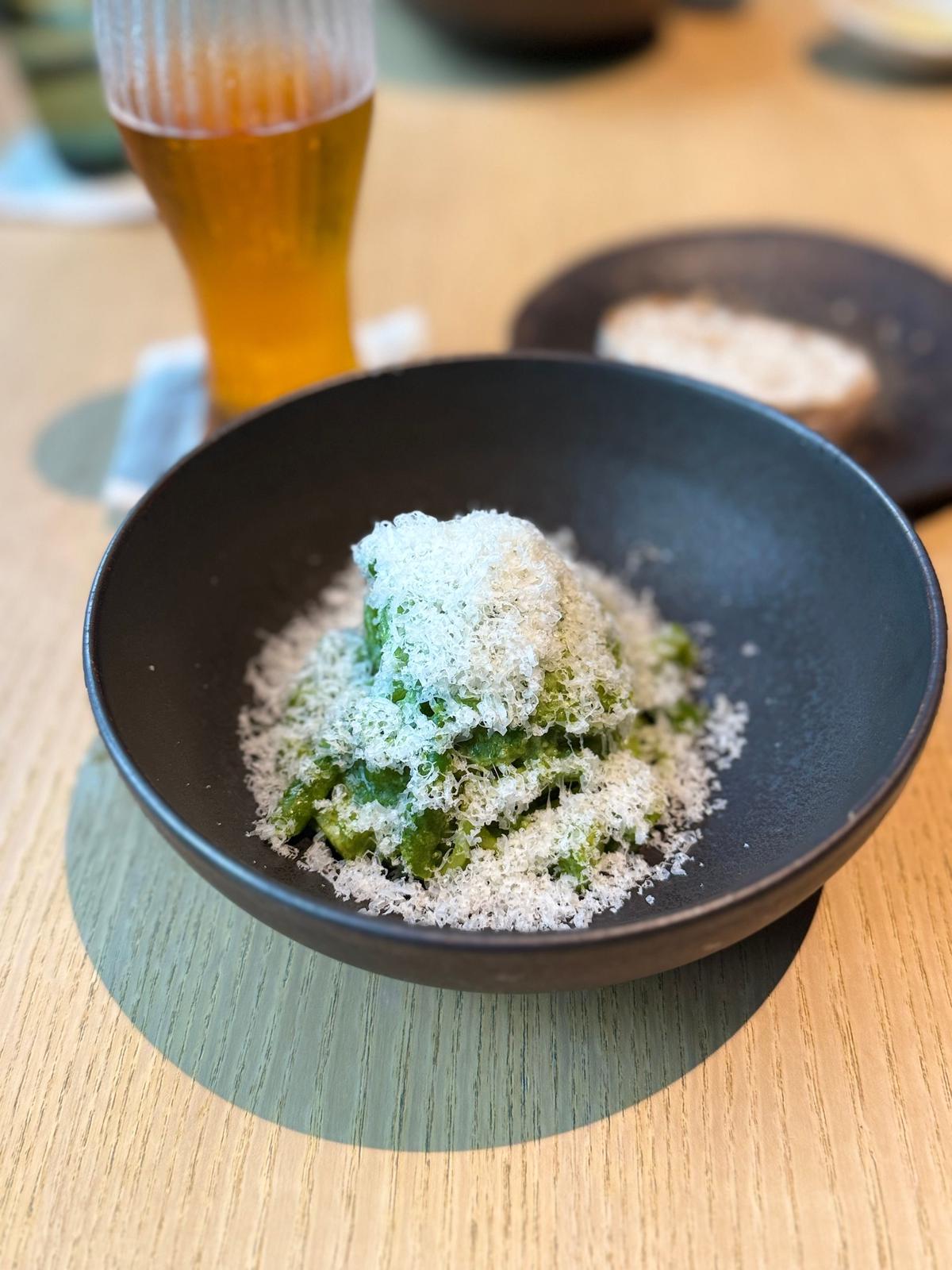 黒い器に入った緑色のペースト状の料理。表面には白い粉末がふりかけられている。背景にはビールとパンが置かれ、木製のテーブルに配置されている。