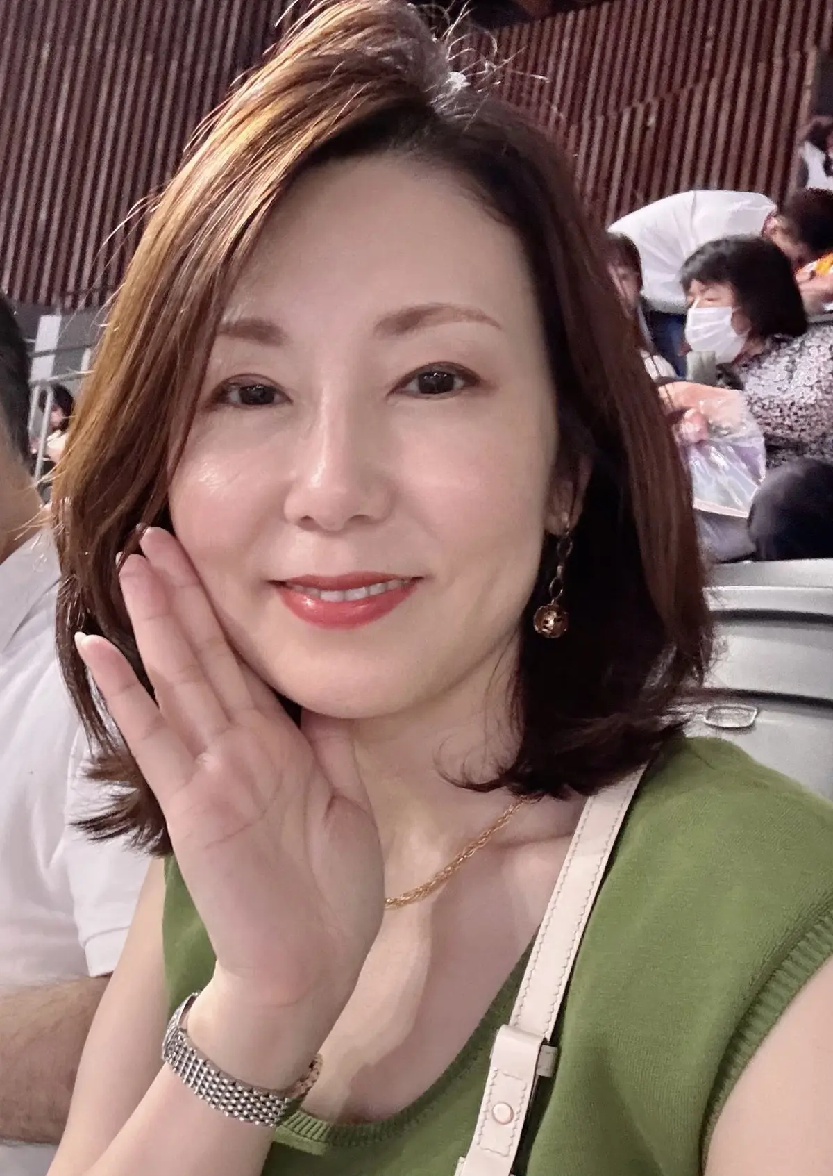 女性が緑のトップスを着て、右手で顔を触えている。耳には金色のイヤリング、首には細いチェーンのネックレスを身につけている。背景には他の人や手荷物が見える。