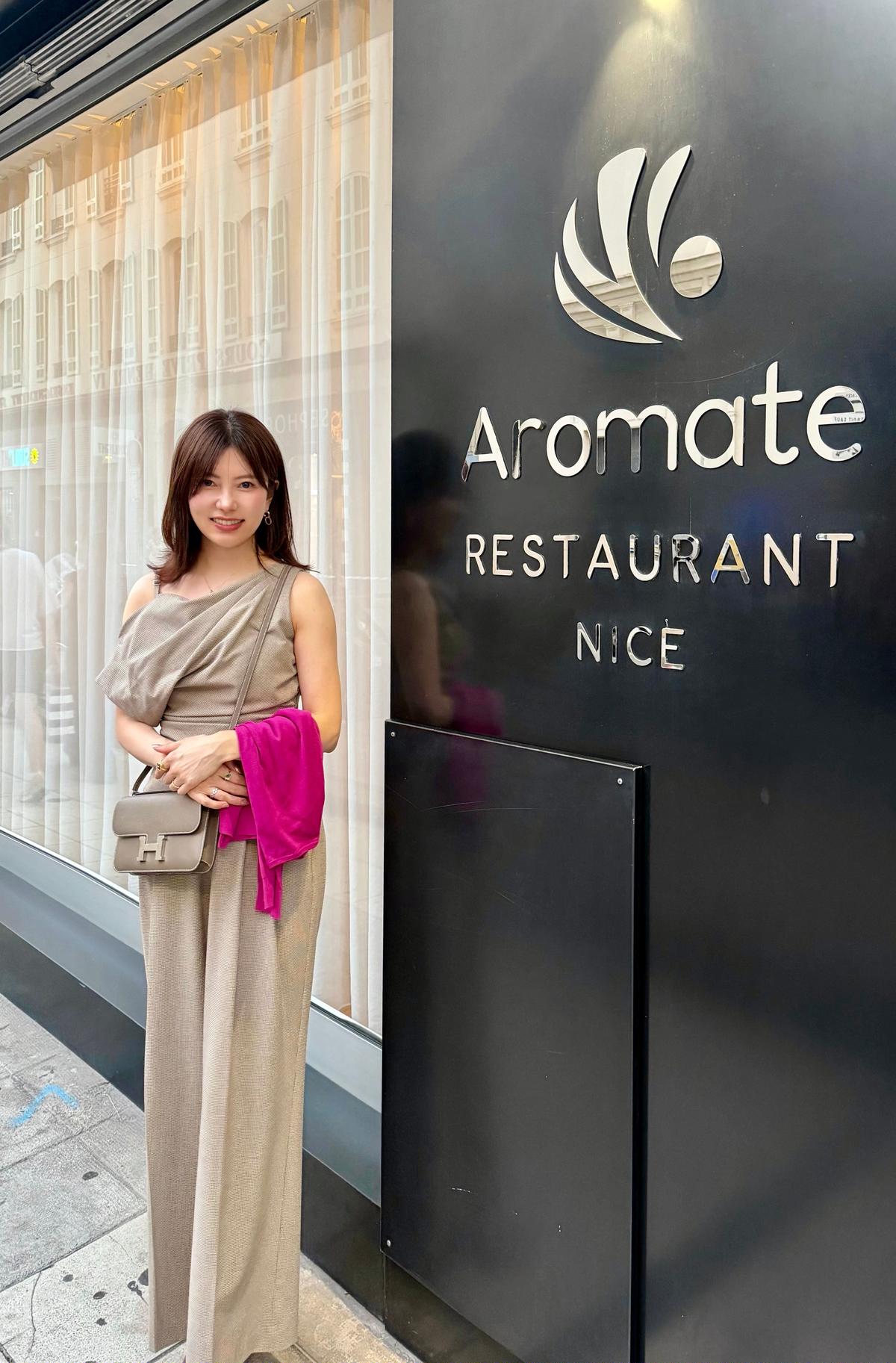 ベージュのロングドレスを着た女性が、レストラン「Aromate Nice」の前で微笑んでいる。右手にピンクのショールを持ち、左腕にはエルメスのバッグを提げている。背景には、建物のショーウィンドウが見える。