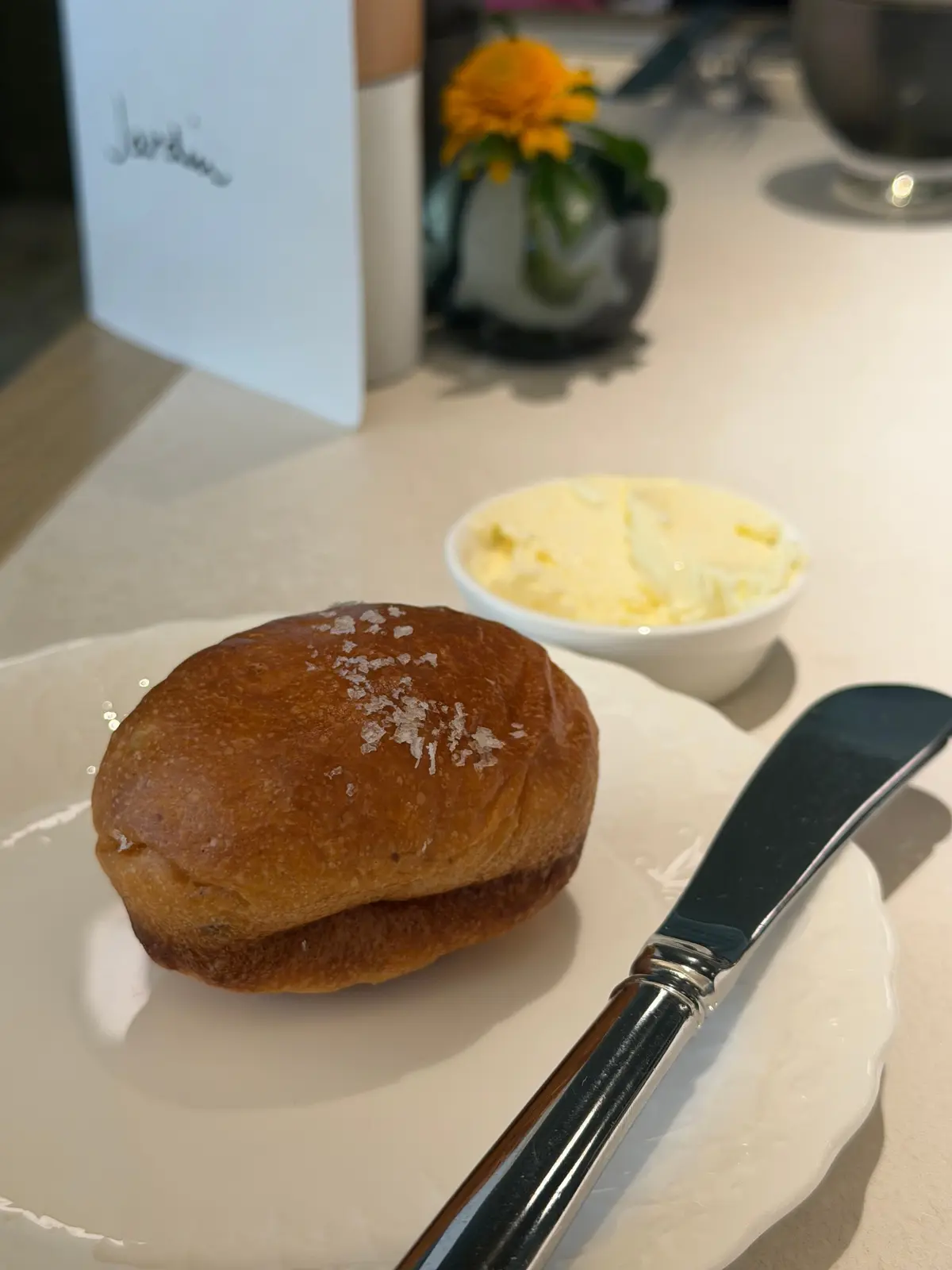 白い皿に置かれた、焼き色のついた丸いパン。パンの上には塩が振りかけられている。皿の横にはバターが添えられ、銀色のナイフが置かれている。背景には、メニューと花瓶がぼやけて見える。
