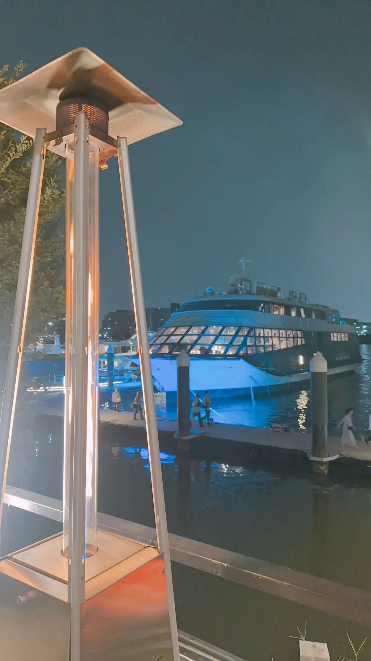 夜の港に停泊した大型客船と、その手前に設置されたパティオヒーター。水面に光が反射し、周囲の建物がぼんやりと見える。