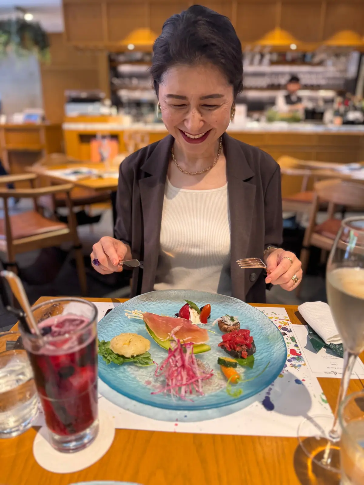 女性が木製のテーブルに座り、青いプレート上の料理をフォークとスプーンで楽しんでいる。プレートには生野菜、魚の切り身、小さな料理が彩り豊かに並べられ、グラスにドリンクが置かれている。背景には明るいカフェの内装と他の客席が見える。