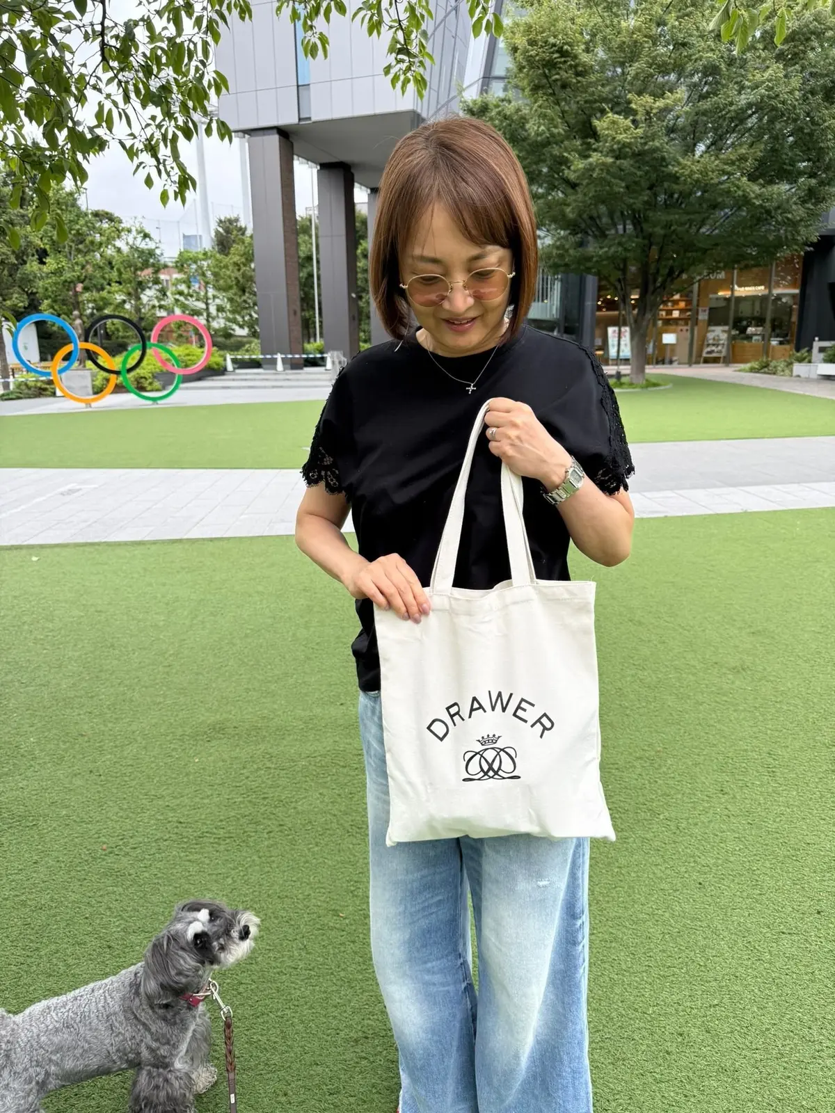 白いトートバッグを持つ女性。茶色のボブヘアに眼鏡をかけ、黒いトップスとジーンズを着用。バッグには「Drawer」のロゴマークが白文字でプリントされている。背景には緑の芝生と建物、そして犬がいる。