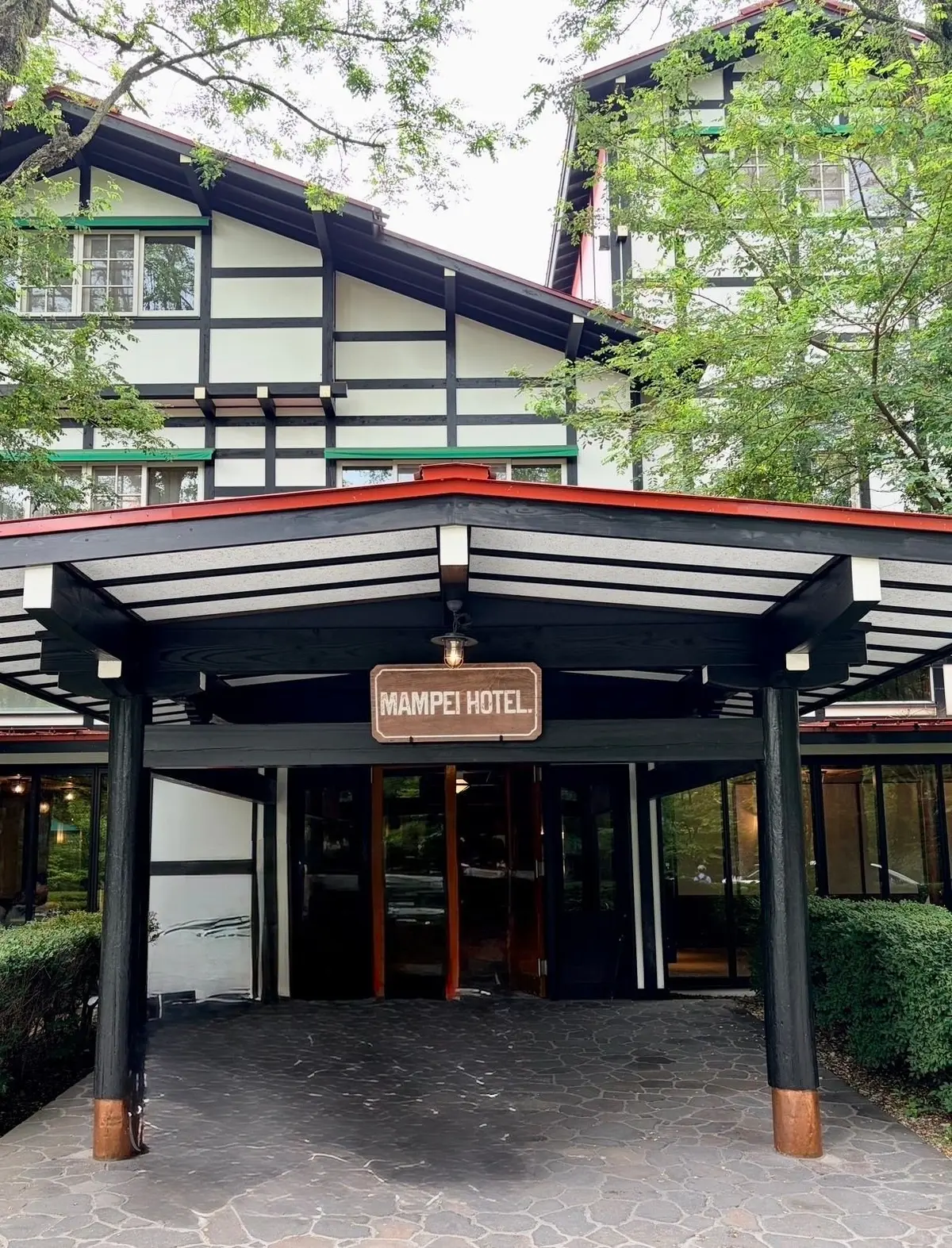 白と黒の木枠とクリーム色の壁が特徴的な建物の入口。屋根付きのポーチがあり、「Mampei Hotel」の看板が掲げられている。石畳の通路がポーチへと続いており、緑豊かな植栽が周囲を取り囲む。