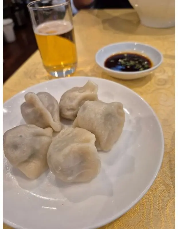 白磁の皿に盛り付けられた、6つの水蒸気を立てる餃子。右隣には醤油ベースの黒いソースが入った白い小皿、さらに右にはグラスに入った淡い色のビールが置かれている。背景は黄色のテーブルクロス。