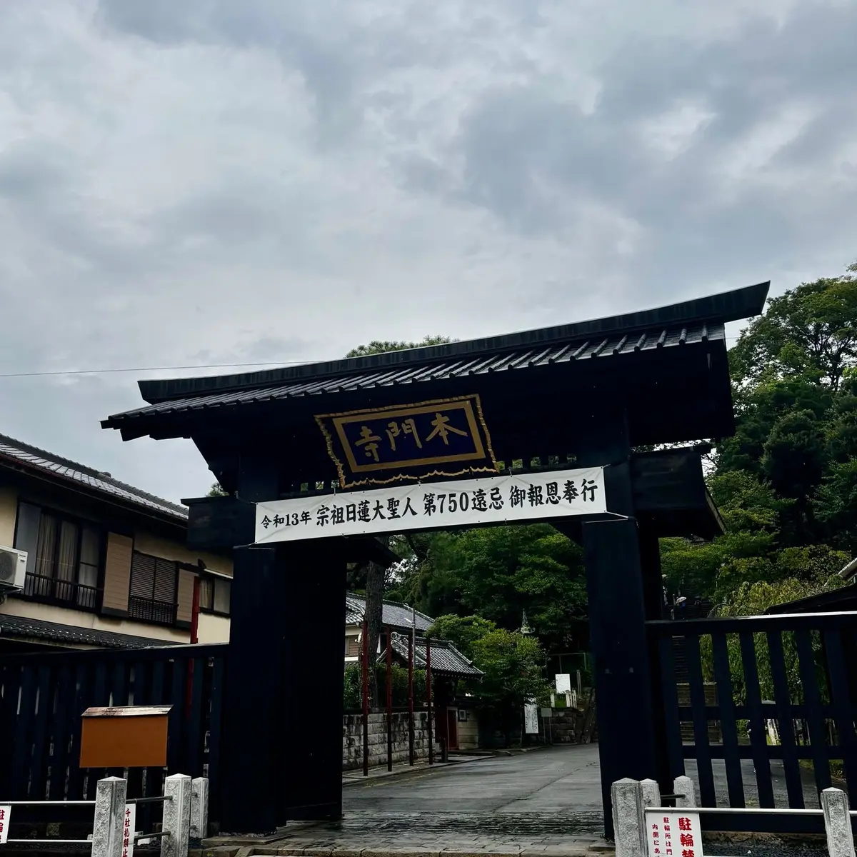 黒い木製の鳥居と、その上にある白い看板。看板には漢字で「寺門木」と書かれ、下には日付と説明文が記載されている。背景には、伝統的な木造建築と緑豊かな木々が見える。曇り空の下、静かな雰囲気。