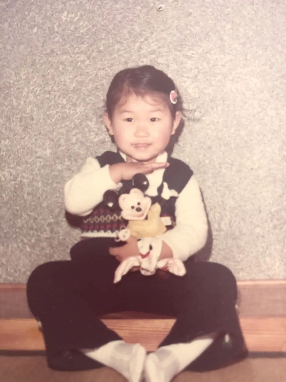 黒いパンツと白い靴下を履いた幼いアジア系の女の子が、ベージュ色のぬいぐるみ人形を抱き、床に座ってこちらを見ている。白い長袖のトップスに黒と白の柄のベストを着ている。背景はグレーの壁。