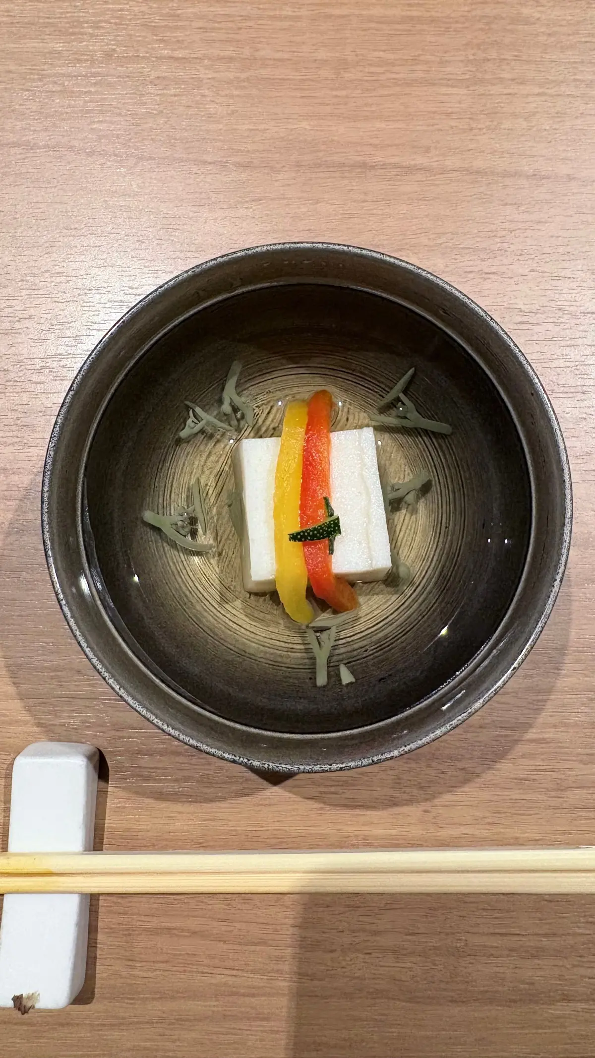 黒い陶器の碗に入った、透明なスープ。白い角状の豆腐と、赤と黄色のパプリカの薄切りが添えられている。碗の底には緑色の葉が散らばり、全体的に上品な盛り付け。