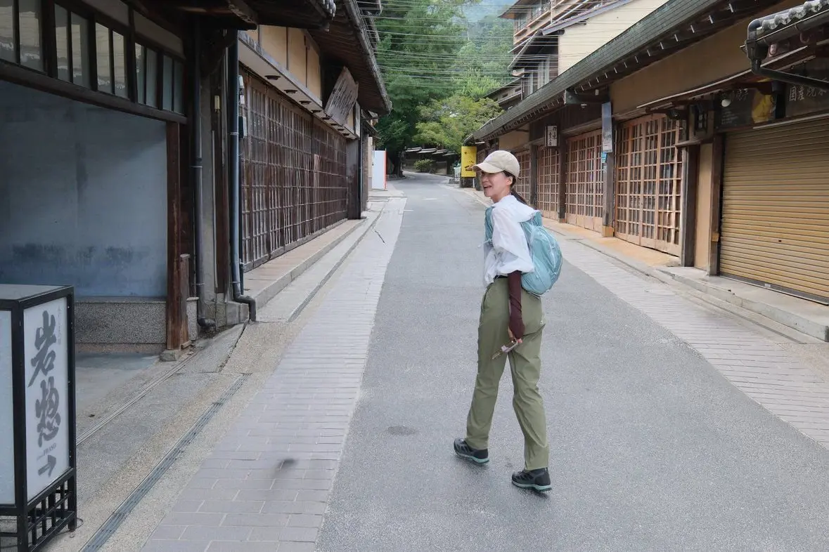 石畳の道が続く並木道。右と左に木造の建物が並び、女性が一人、白の帽子とリュックサックを背負って歩いている。道の脇には黒い看板が立っている。