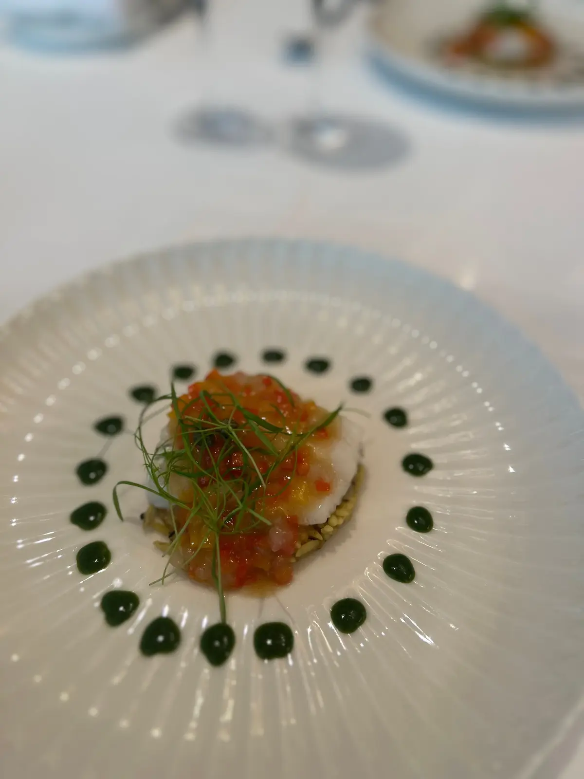白磁の皿に盛り付けられた、魚料理の一皿。中央には白身魚が乗っており、その上には赤色の魚卵と緑色のハーブが散らされている。皿の周りには緑色のソースが点状に配置されている。