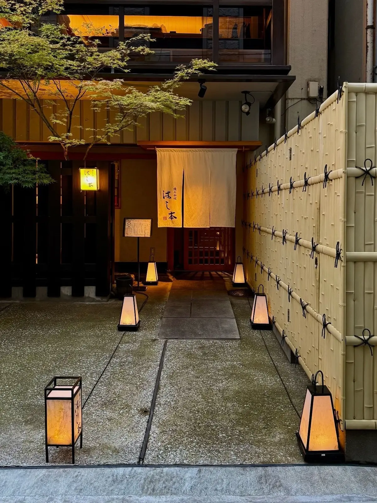 日本の飲食店への入り口。砂利敷きの通路に、和風の灯籠が並んでいる。右側には竹製の仕切りがあり、建物は暖色系の照明で照らされている。