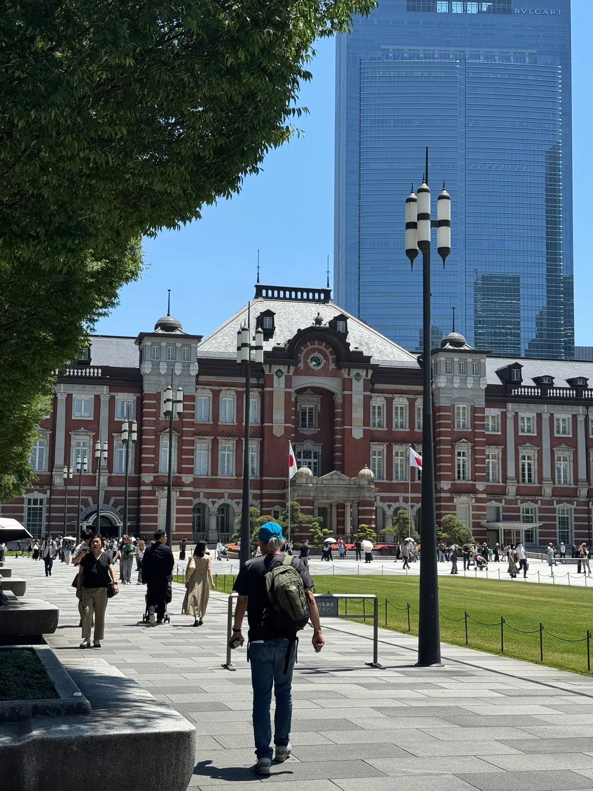 東京の広場に人々が歩く。赤レンガの歴史的建物とモダンな高層ビルが背景にあり、木々や街灯が並ぶ。