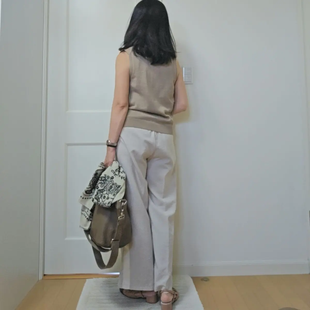 ベージュのタンクトップとワイドパンツを着用した女性が、ドアの前に立っている全身写真。右手に茶色のバッグを持ち、左腕には柄入りのスカーフが巻かれている。背景は白の壁。