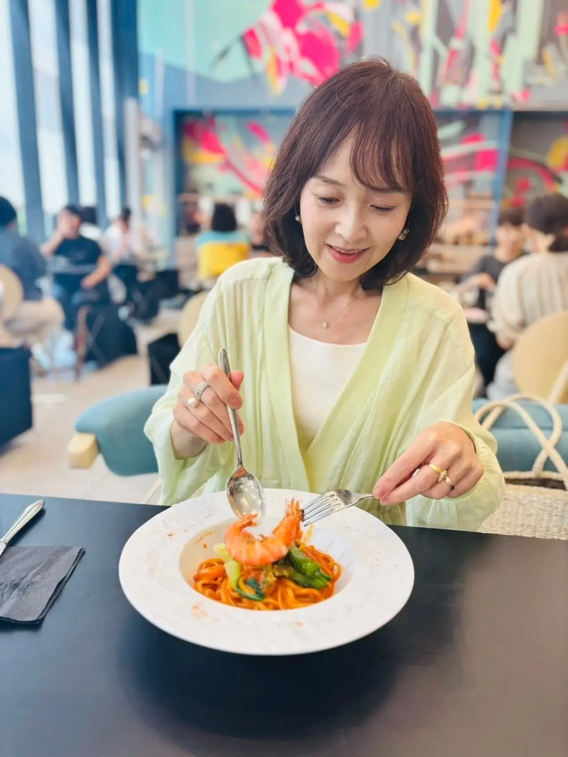 レストランでパスタを食べる女性。茶色のボブヘアで、緑色のカーディガンを羽織っている。白い皿に盛り付けられたパスタには、エビや野菜が添えられている。背景には、カラフルな絵画が飾られた空間。