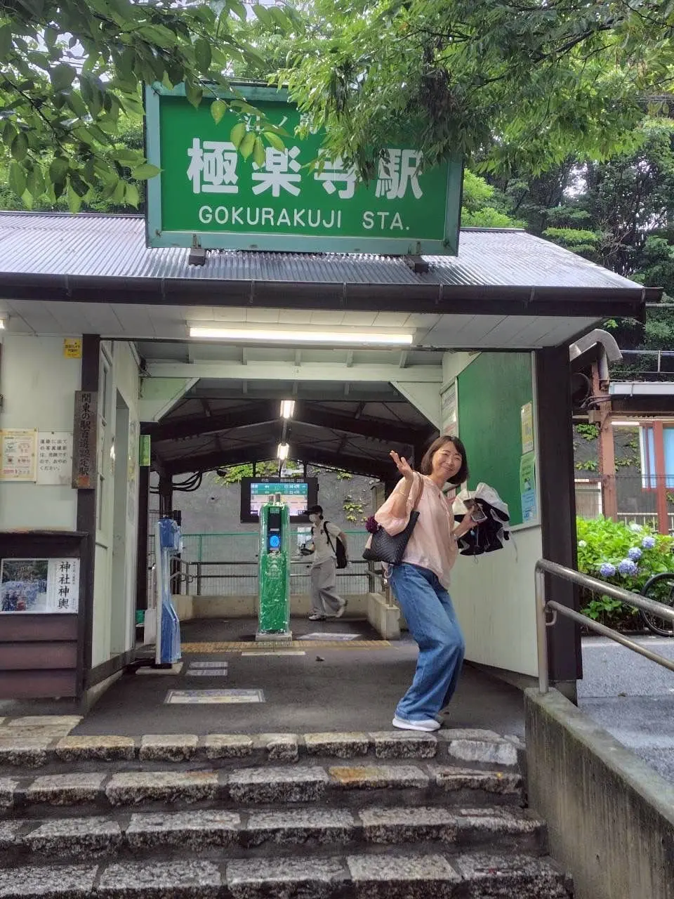 御蔵庫寺駅前の石段に立つ女性が片手を上げてカメラに向かって波手を打っている。女性はピンクのトップスとジーンズを着用し、黒のショルダーバッグを肩にかけている。背景には緑の駅看板と駅舎があり、別の人が駅の方向に向かって歩いている。