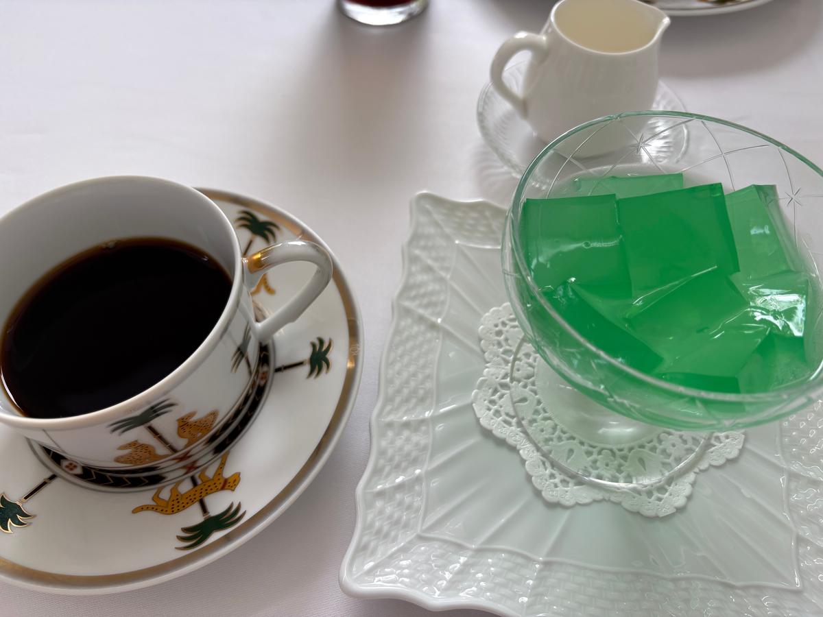 白テーブルに置かれたコーヒーカップとゼリー。カップは白地に金と茶色の装飾が施され、ゼリーは透明なグラスに盛り付けられ、鮮やかな緑色をしている。