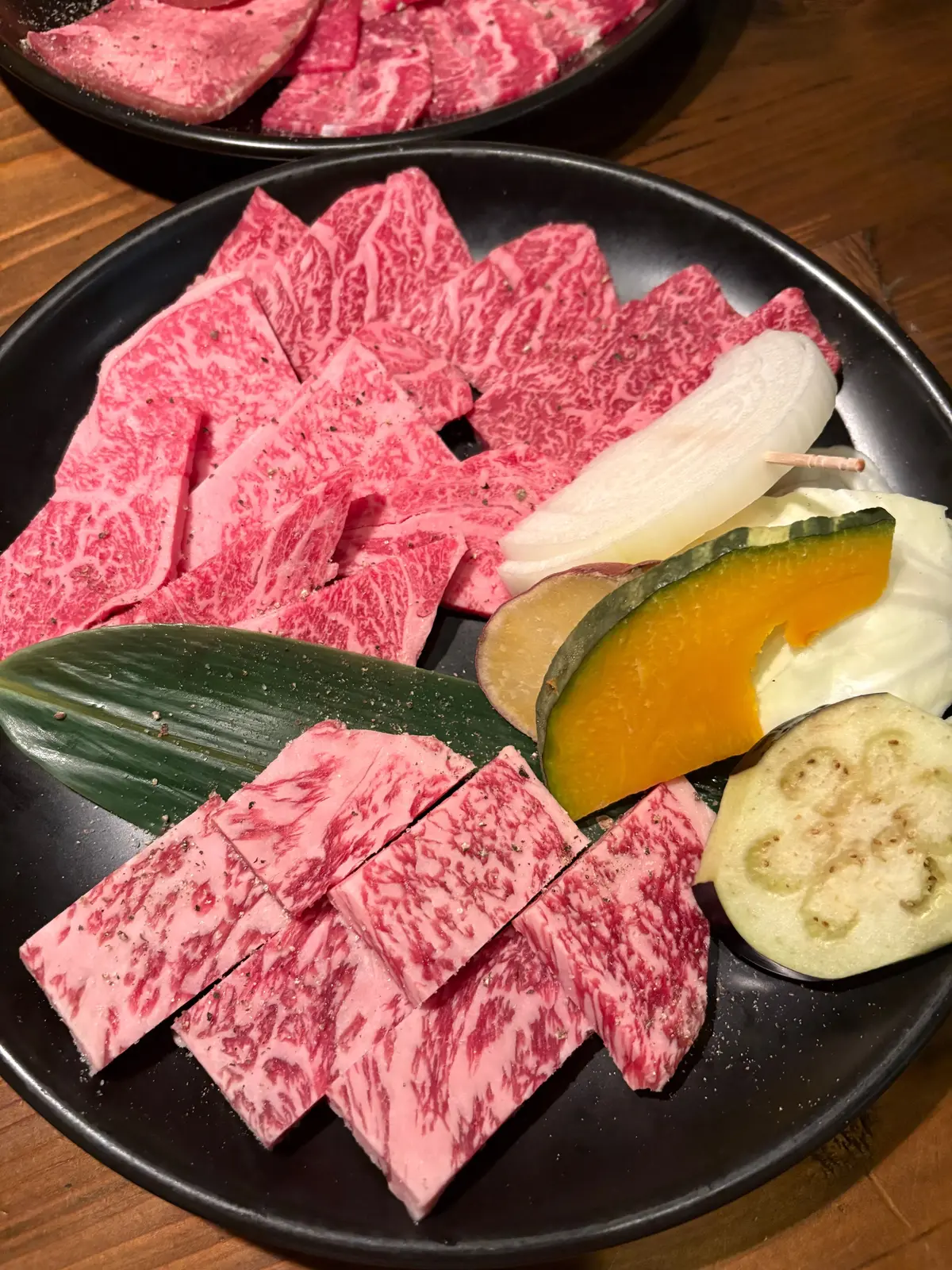 黒い皿に赤身の牛肉の切り身が並び、緑の葉、黄色い果物、薄い緑の野菜が添えられている