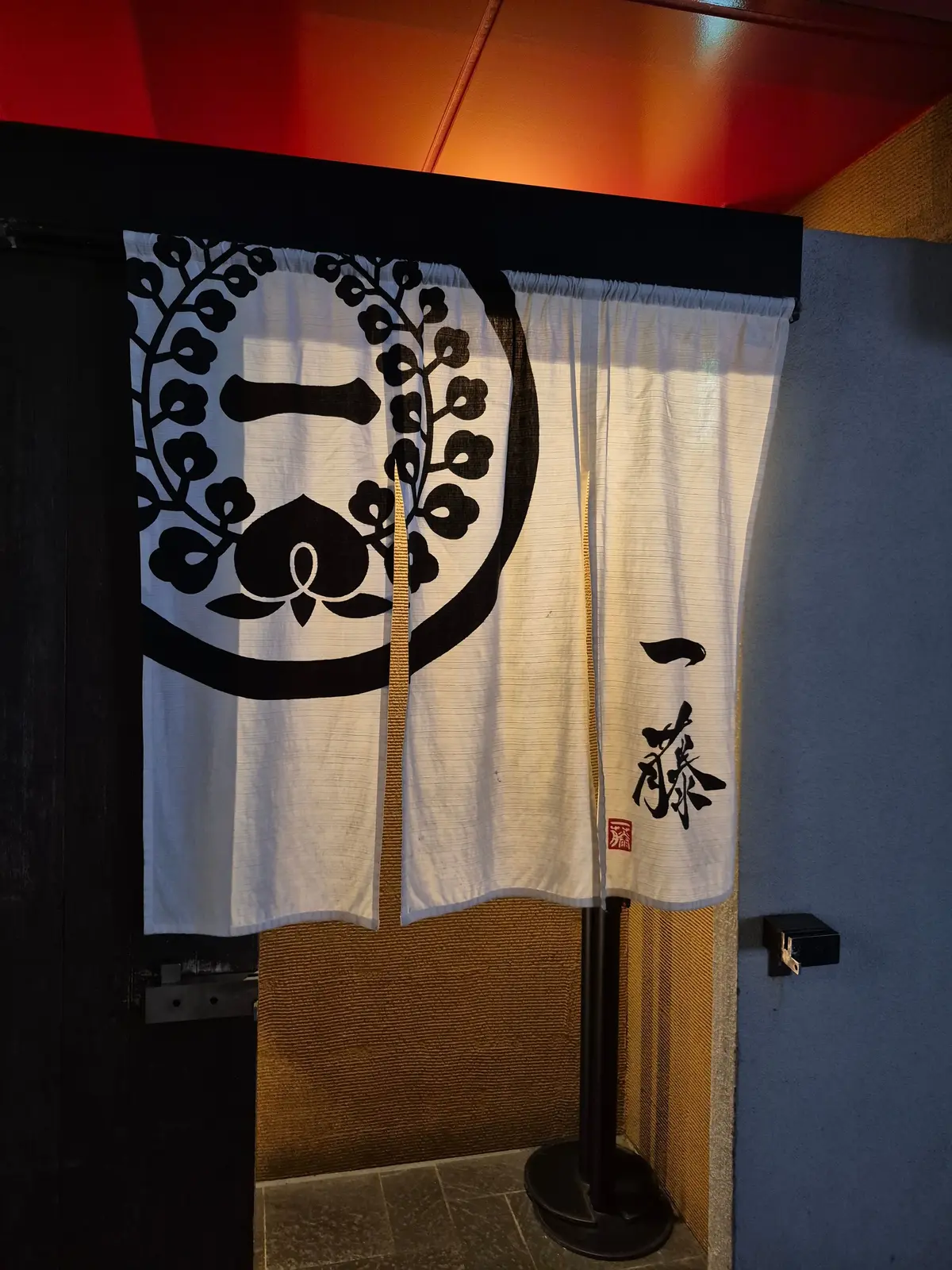 白地ののれん。左側には黒い丸の中に植物の模様、右下には赤い文字で「一茶」と書かれている。黒い扉の前に設置され、下には藁のマットが敷かれている。