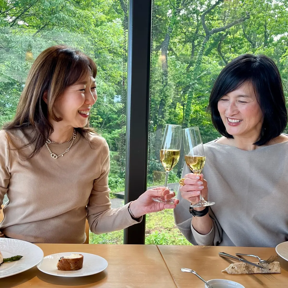 二人の女性が窓のそばのテーブルに座り、ワイングラスを掲げて笑っている。テーブルには料理と飲み物が置かれている。