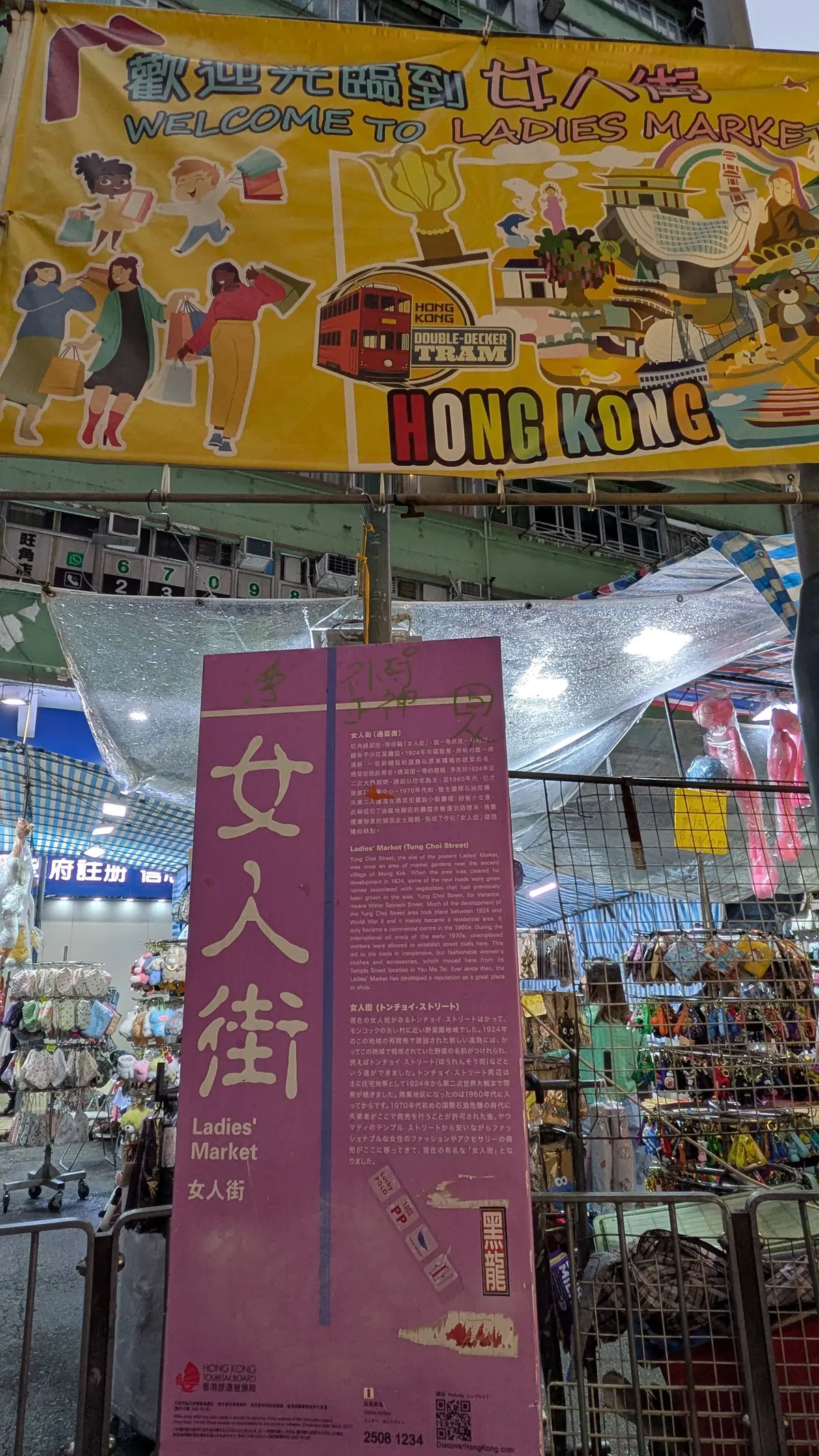 鮮やかな黄色のバナーが「Ladies' Marketへようこそ」と英語と中国語で書かれ、香港の観光スポットを案内している。背景にはトラムや市場のイラストが描かれている。