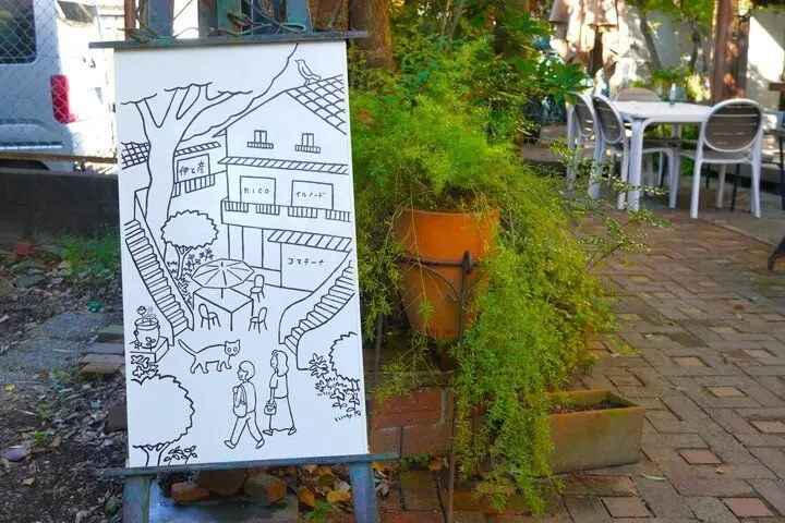 手書き風のイラストが描かれた看板が、植栽のそばに立てられている。看板には、建物や人物、猫などが線画で表現されている。背景には、テーブルと椅子が並んだテラス席が見える。
